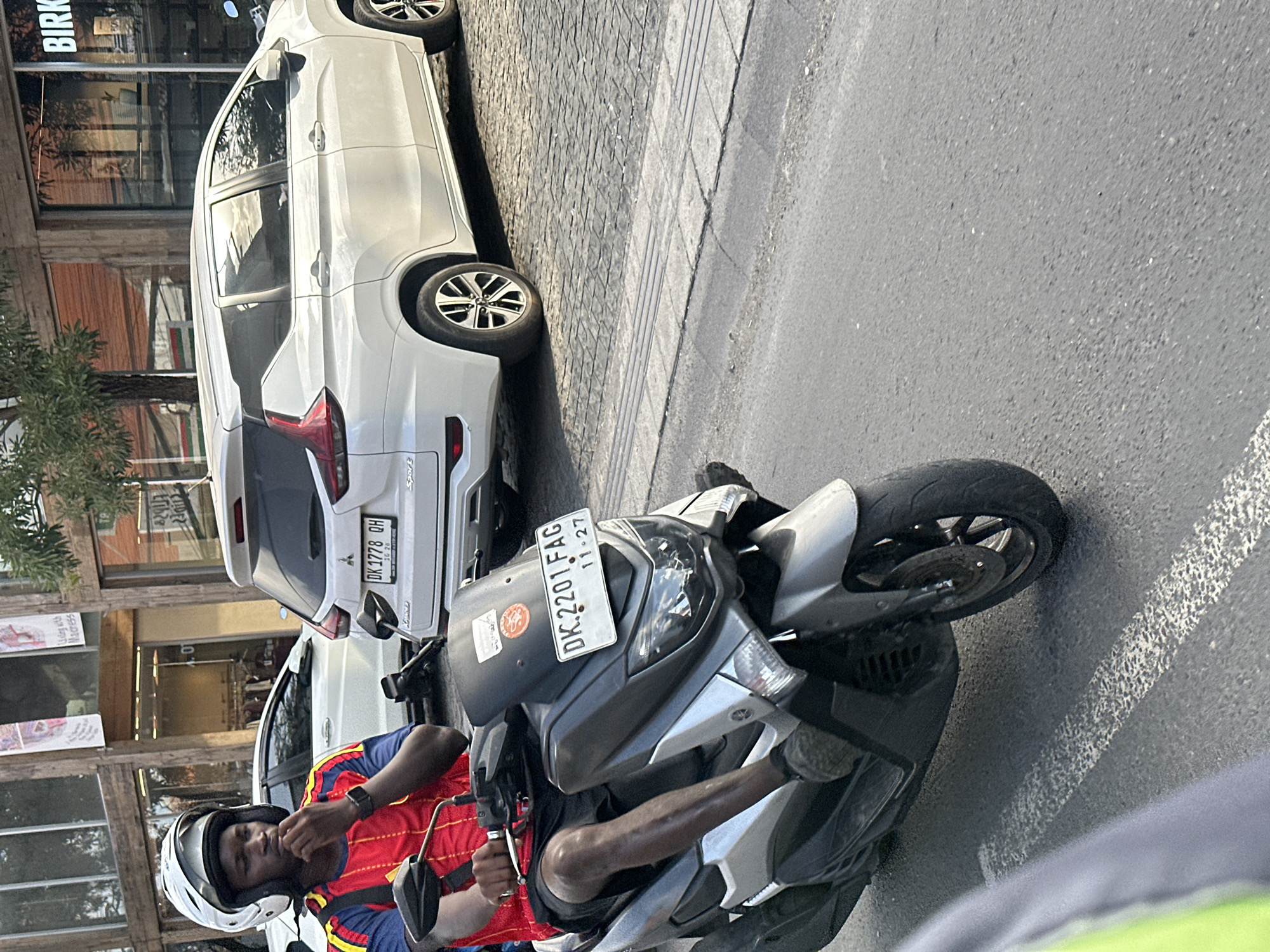 A man wearing a white helmet and a red, yellow, and blue shirt is riding a silver and black scooter on a paved road. He is looking to his left and has his right hand on the handlebars. The scooter has a license plate that reads "DK.2201 FAG 11.27". To the right of the scooter is a white car with a license plate that reads "DK 1778 OH". In the background are buildings with shops, including one with the word "BIRK" on it. The environment appears to be a street in North Kuta, Indonesia.