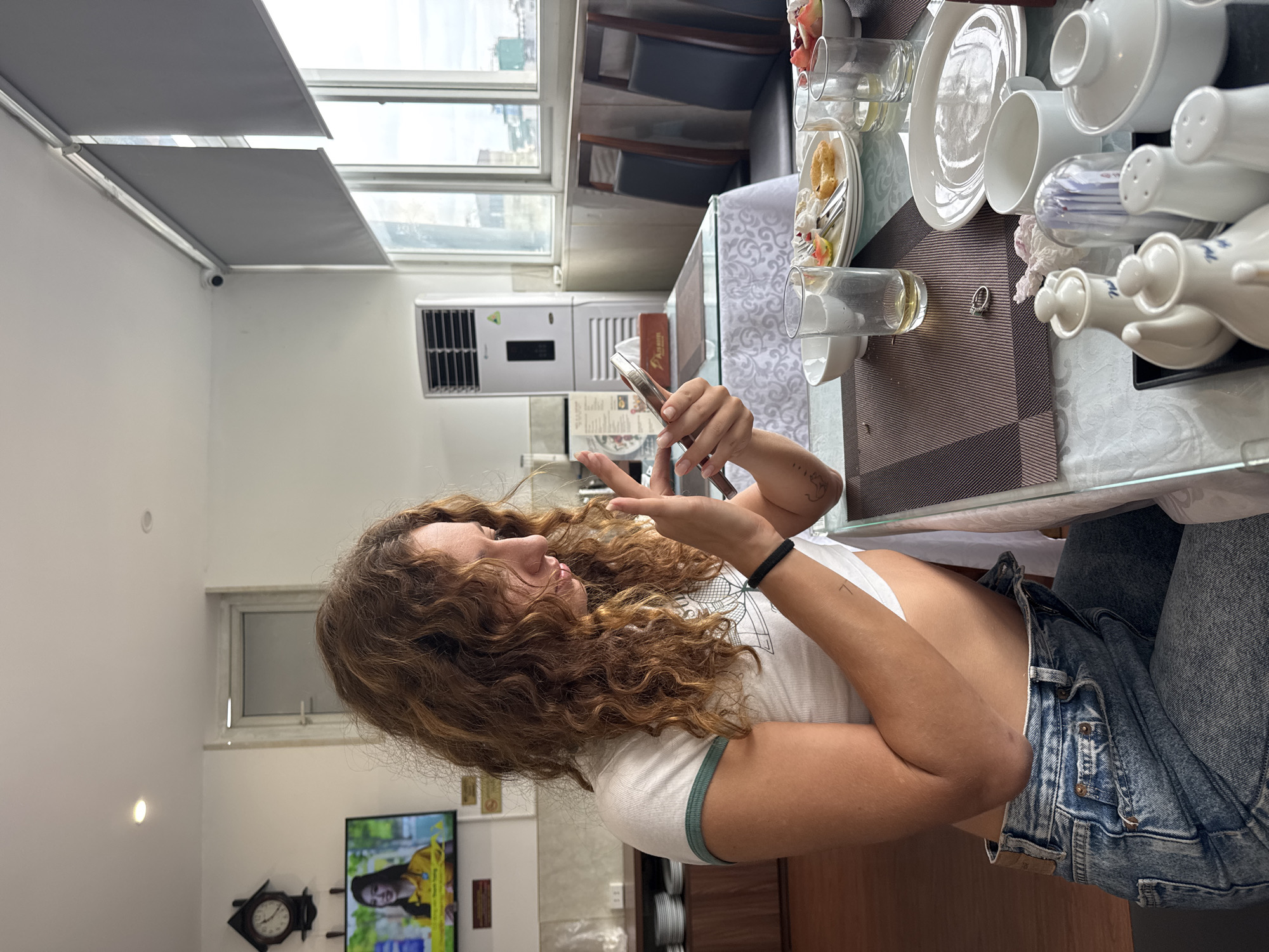 A young woman with curly brown hair is seated at a table in a hotel dining area in Hanoi, Vietnam. She is wearing a white crop top with green trim and blue denim jeans. She is looking down at her phone, which she holds in her hands. The table is set for breakfast, with plates of food, glasses, and cutlery. In the background, there is a television screen displaying an advertisement, a clock, a large white air conditioning unit, and windows with a view of the city. The dining area appears to be part of a hotel, indicated by the decor and amenities.
