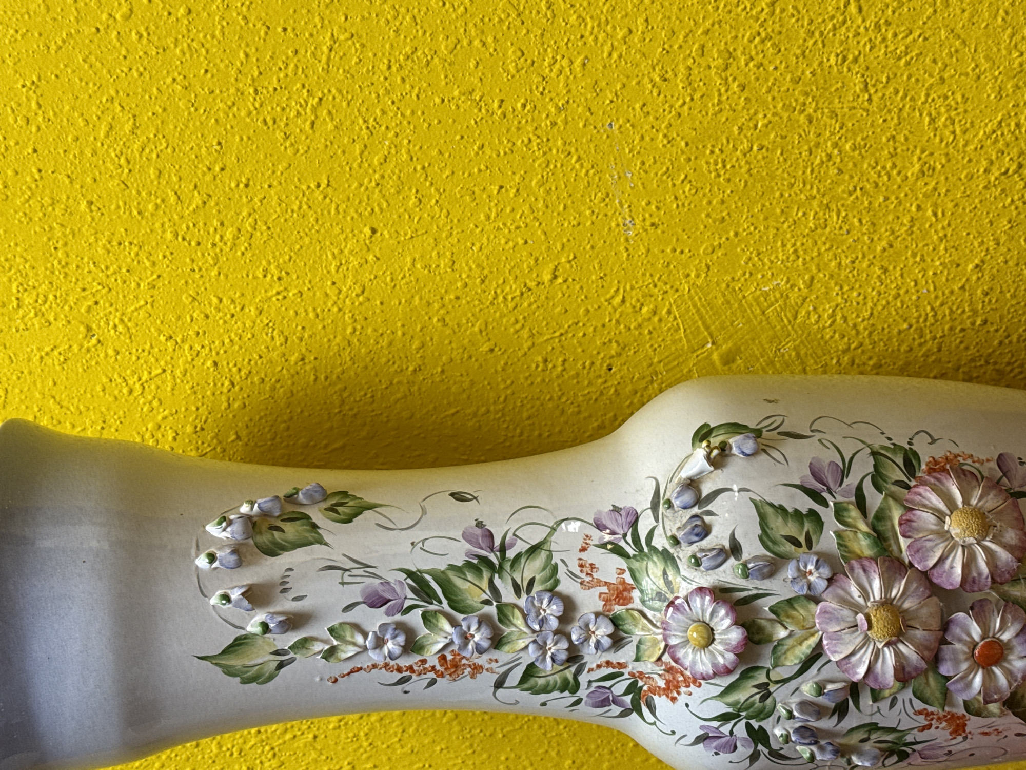 The image shows a decorative white vase with a floral design, resting against a bright yellow textured wall. The floral design on the vase includes purple, white, and green elements. The texture of the yellow wall is granular and rough.