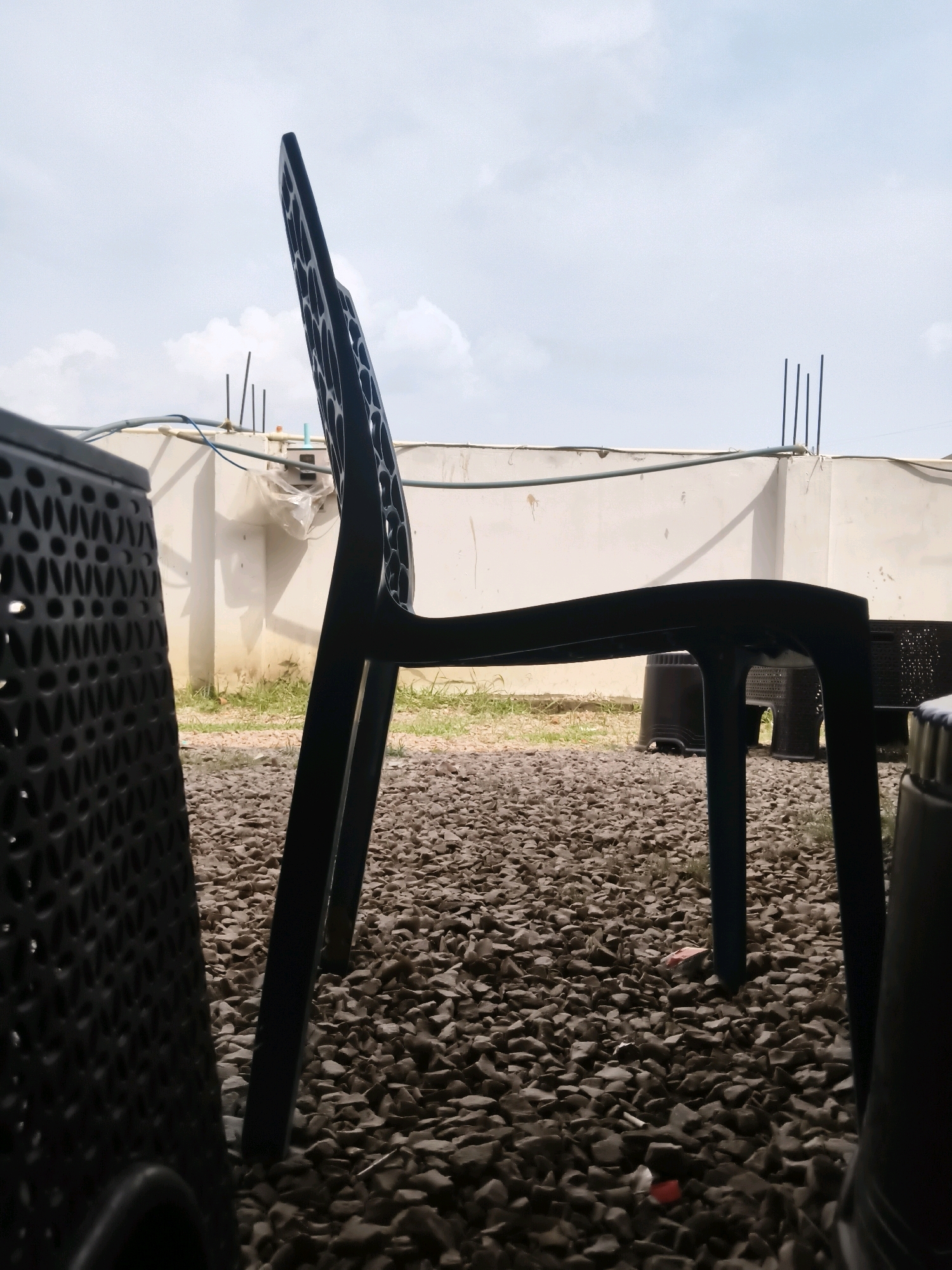 The image depicts an outdoor scene with a focus on a black plastic chair positioned in the foreground. The chair is seen from a low angle, with its back prominently displayed, featuring a perforated design. To the left and slightly behind the main chair, another black plastic object, possibly a chair or table, is visible, also with a perforated texture. The ground is covered in small, dark-colored gravel. In the background, a light-colored wall or barrier runs horizontally, with some pipes and electrical wires attached to it. Above the wall, there are visible rebar structures protruding, suggesting ongoing construction. The sky is overcast with clouds. The location is identified as Goondpur, India. No people are present in the image.