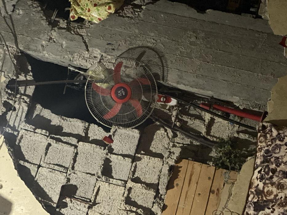 A room in disrepair features a collapsed ceiling, revealing exposed cinder blocks and rebar. An oscillating fan with red blades and a black cage stands near the damaged area. Electrical wires and a red pipe run along the deteriorating wall. Wood planks and part of a floral-patterned cushion suggest the room is in use, despite its dilapidated state. A small potted plant sits near the base of the wall.