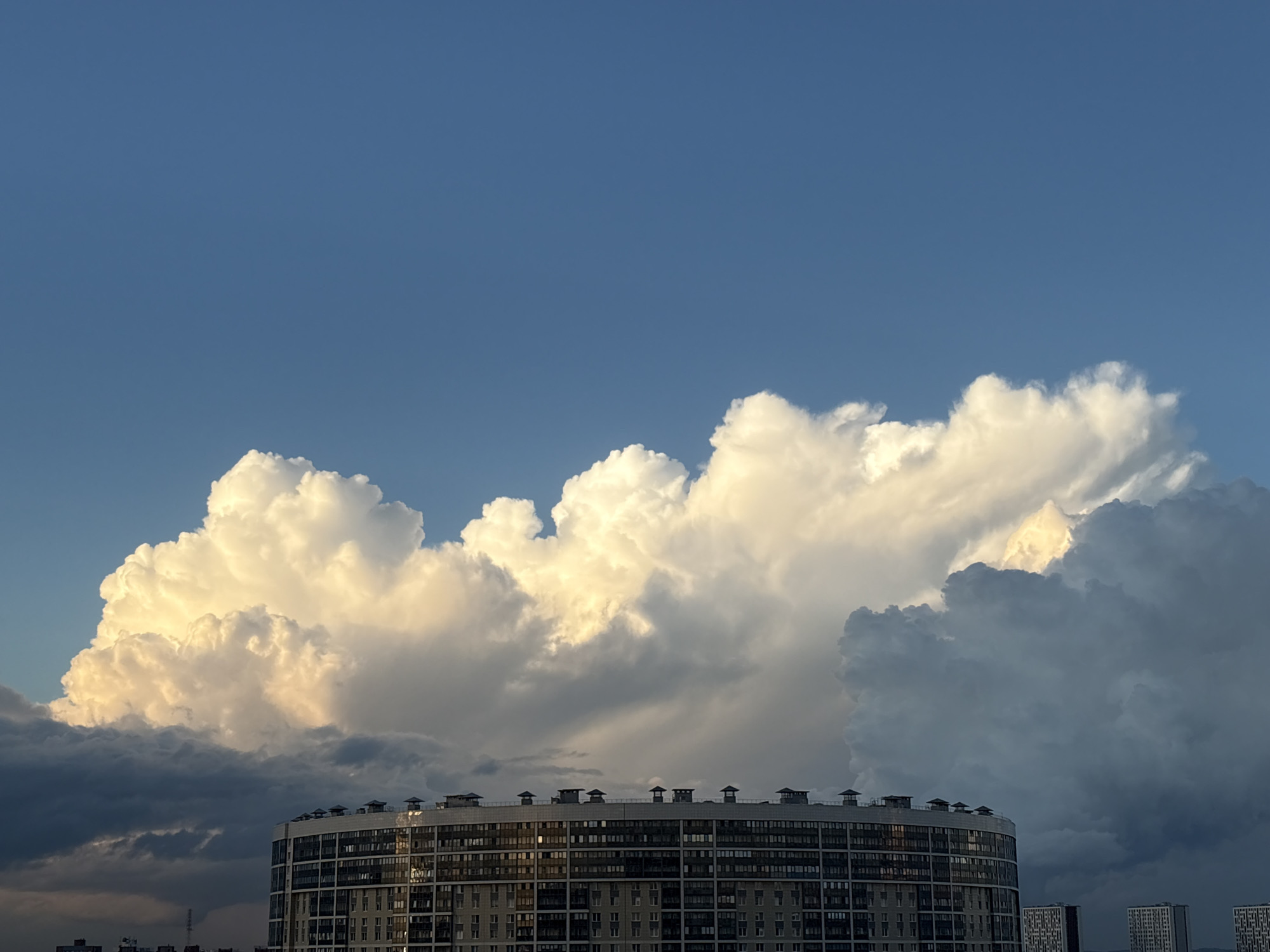 A wide shot captures the sky above St. Petersburg, Russia, dominated by a dramatic display of cumulus clouds. The sky is a clear, deep blue, transitioning to a lighter hue towards the horizon. A large, luminous cumulus cloud formation, brightly lit from within by the sun, takes center stage in the middle ground. It is flanked by darker, more ominous-looking clouds on either side, suggesting an approaching storm or the remnants of one.
In the foreground, a curved, multi-story residential building with a modern facade occupies the lower third of the frame. The building is characterized by numerous windows, a light-colored exterior, and a row of small, dark structures on its roof, possibly ventilation shafts or decorative elements. The building's rounded shape is evident, curving slightly towards the right side of the image. In the background, behind the main building, two smaller, rectangular residential buildings are visible, their facades also featuring many windows. The overall scene is one of natural grandeur set against an urban backdrop. There are no people visible in the image.