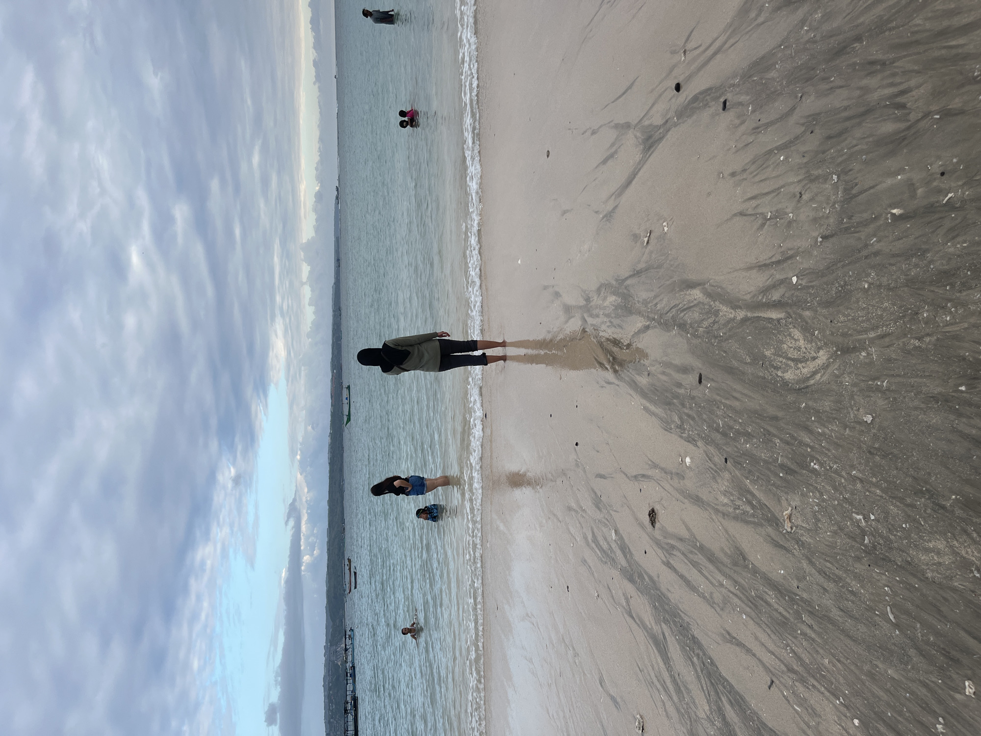 A vertical, eye-level shot captures a wide view of a beach in Kuta, Indonesia, under a cloudy sky. The foreground is dominated by wet, sandy beach with dark sand streaks that have been washed in by the tide. In the mid-ground, the turquoise ocean meets the shore with gentle waves. Several people are visible in the shallow water. On the left, two children are playing in the water, one partially submerged and the other fully. To the right of them, a woman in a black shirt and denim shorts stands in the water, facing the ocean. Further to the right, a woman wearing a hijab and dark pants is standing on the wet sand, facing away from the camera. In the background, several small boats are moored in the calm ocean, and a distant shoreline with buildings is visible. On the far right, a lone individual stands in the water. The overall scene depicts a tranquil beach atmosphere with people enjoying the water.