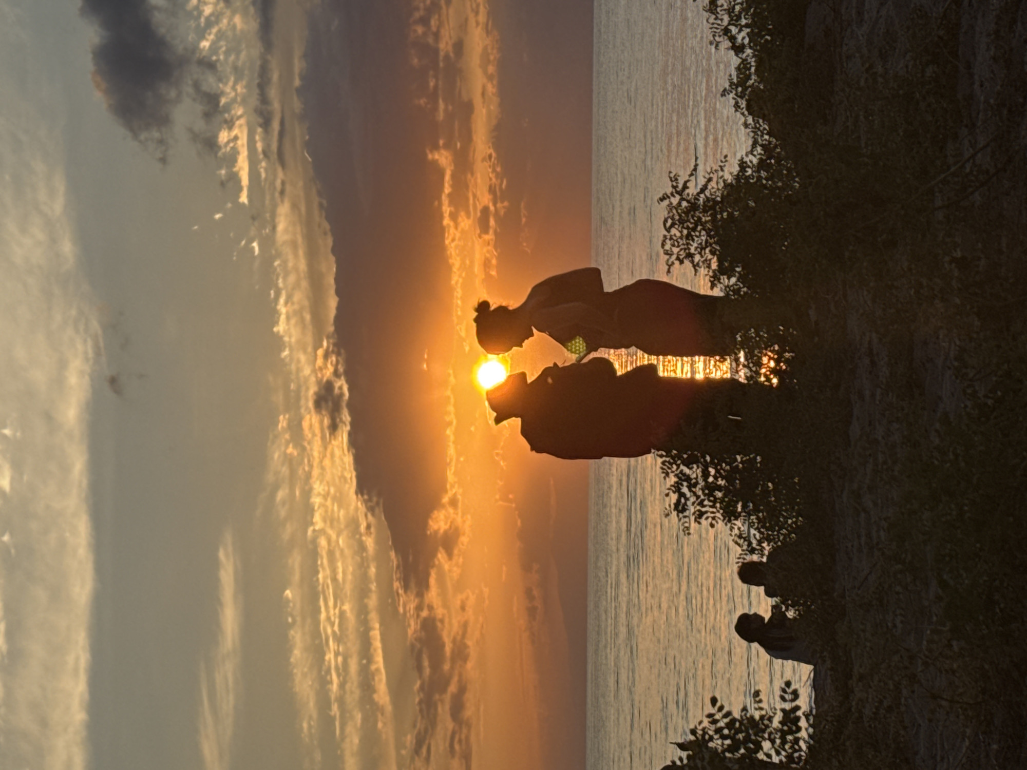 The image captures a sunset over the Issyk-Kul lake near Basbeshik, Kyrgyzstan. Silhouettes of people are visible on the shore, with two individuals standing prominently in the foreground, facing each other. The sun is setting, casting a warm orange glow across the sky and reflecting on the water. Clouds are scattered in the sky, adding texture to the sunset. In the background, a faint outline of mountains can be seen on the horizon. The foreground is dark, with silhouettes of trees and foliage obscuring the lower part of the scene.