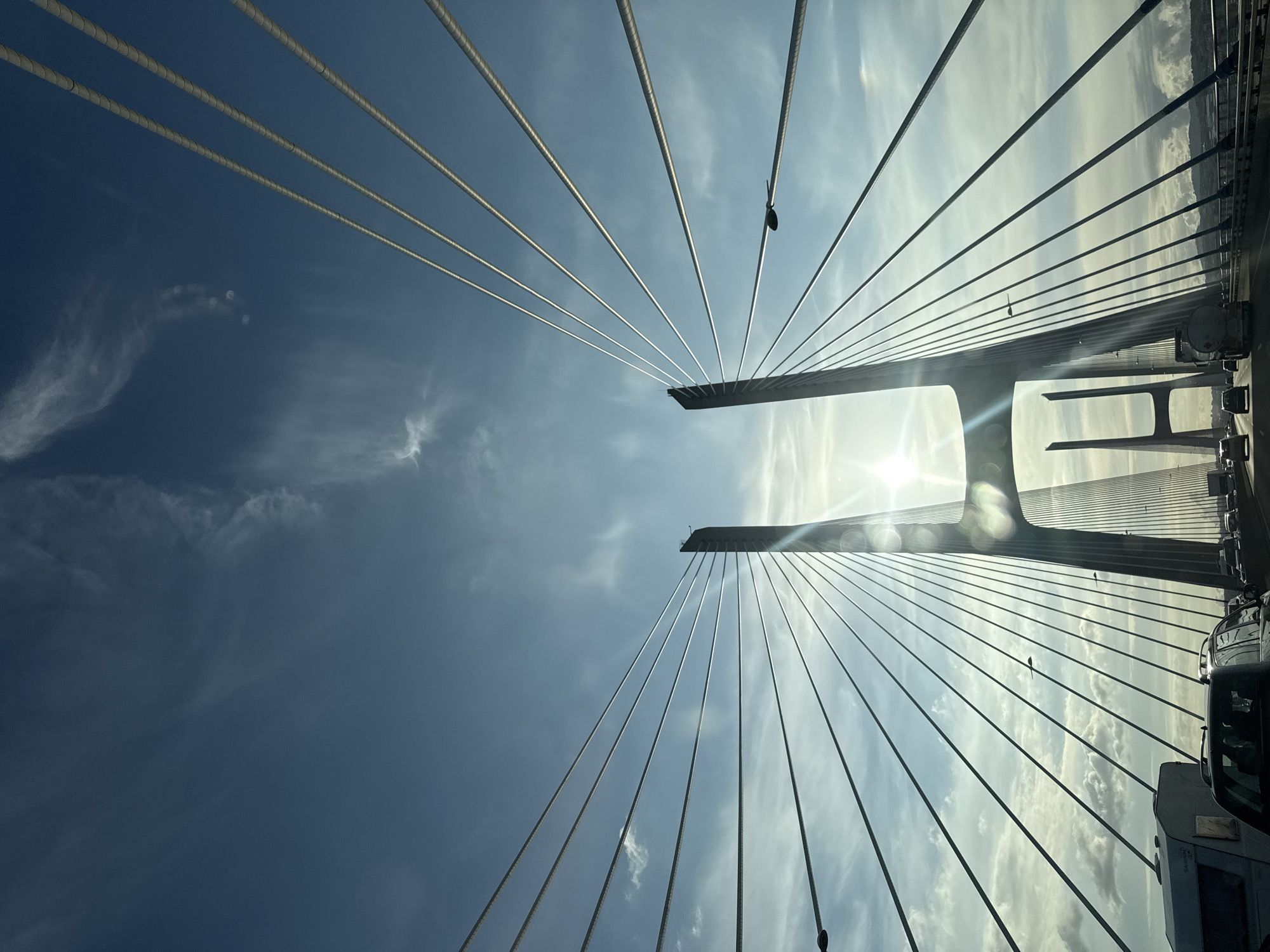 An upward-facing, low-angle shot captures the grandeur of a cable-stayed bridge against a bright, partly cloudy sky. The central elements are the numerous steel cables, fanning out in a symmetrical pattern from two prominent, dark pylon-like structures positioned in the middle distance. These structures are partially silhouetted against the intense glare of the sun, which is situated behind them on the right side of the image, causing a significant lens flare and bright reflections.
The sky is predominantly clear blue, with scattered wispy white clouds adding texture. The bright sunlight suggests it is daytime, possibly morning or late afternoon. On the far right edge of the frame, the tops of several vehicles, including cars and a truck, are visible, indicating ongoing traffic on the bridge deck. A small, dark bird is perched distinctly on one of the tensioned cables on the right side of the composition.
The perspective emphasizes the converging lines of the cables, creating a sense of immense scale and architectural design. Given the provided location context of Barreiro e Lavradio, Portugal, this scene likely depicts a section of the Vasco da Gama Bridge. No visible text is present in the image.