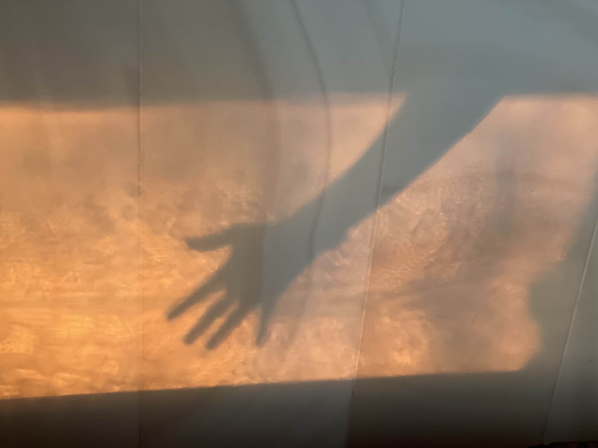 A photograph shows a shadow of a hand projected onto a wall, illuminated by an orange light. The wall appears to be made of light-colored panels. The light source appears to be from outside, possibly sunlight at sunrise or sunset. No specific location details within Riga, Latvia, can be determined from the image. No other people, subjects, or objects can be discerned.