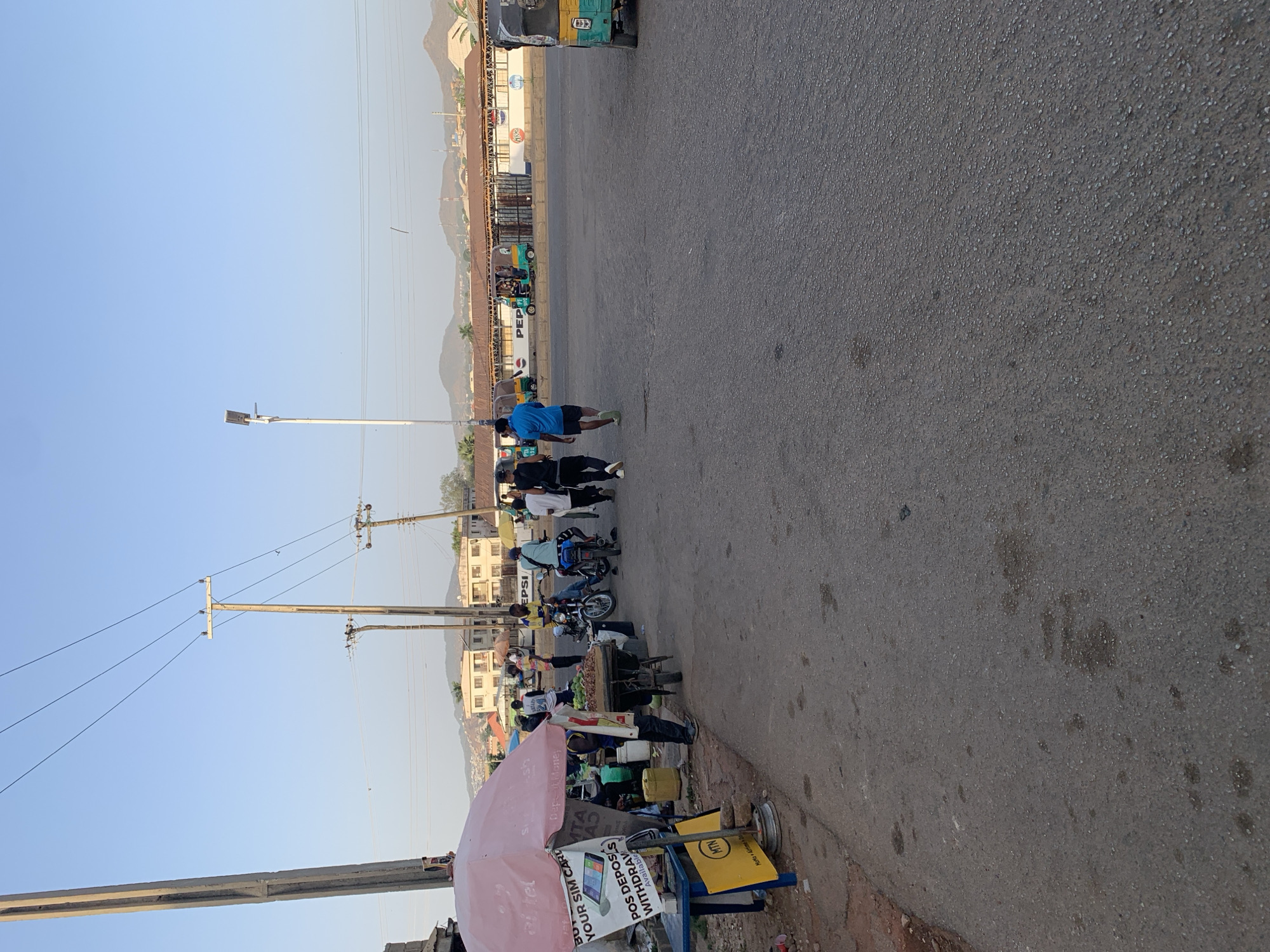 The image captures a street scene near New Stadium, Nigeria. Along the street, there's a line of people, some possibly interacting with vendors or merchants on the roadside, several motorcycles are parked nearby, and there's a person jogging in a blue shirt and black shorts, There is what appears to be roadside vending activity with items displayed for sale, and an MTN-branded umbrella covers part of the vending setup. Power lines and poles are visible overhead, and in the background, one can see buildings, hills, and mountains. A vehicle is approaching.