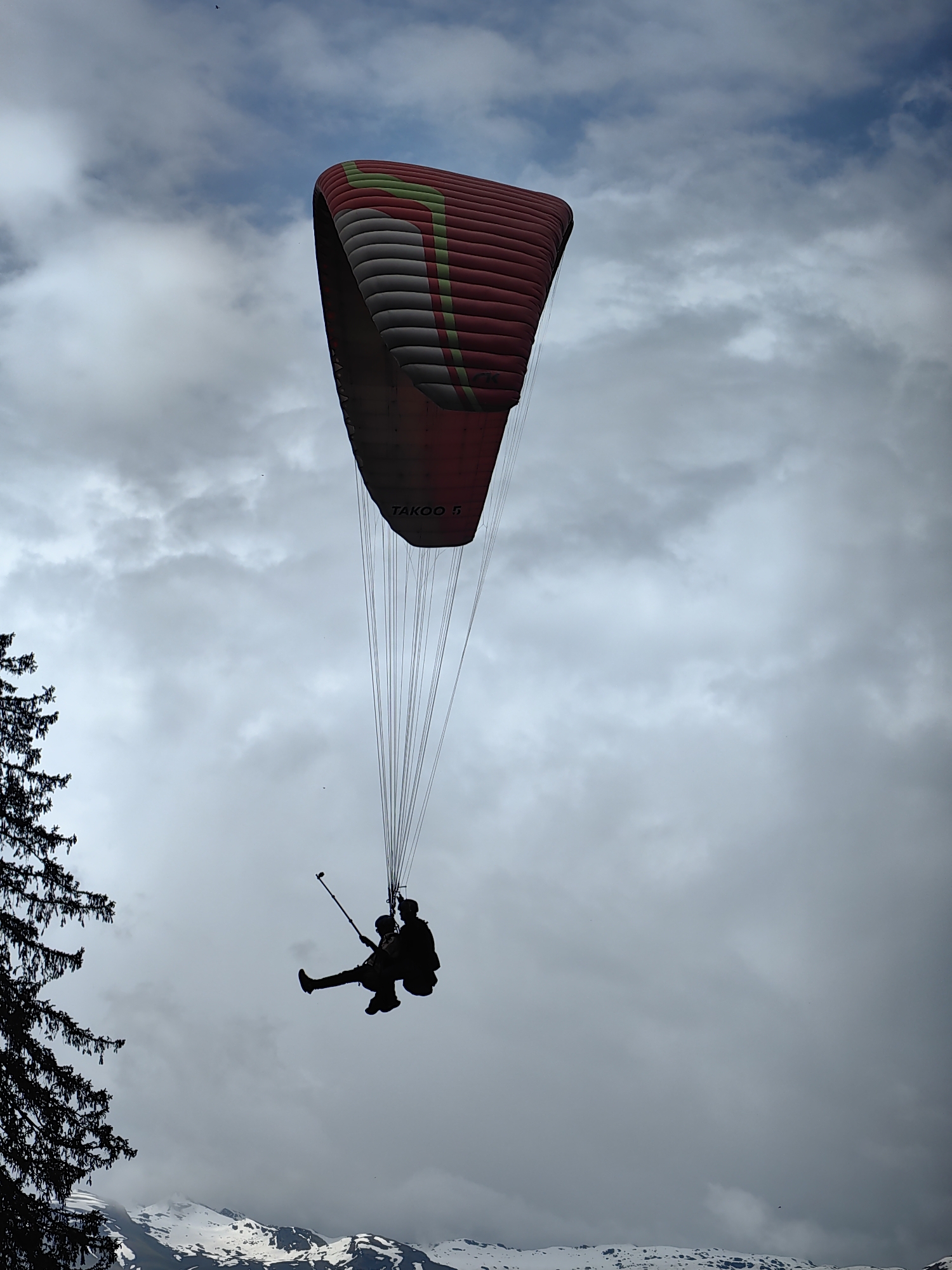 This media file captures a tandem paragliding experience in Solang, India. The main subjects are two people silhouetted against a cloudy sky, suspended beneath a large, red, grey, and green paraglider. The paraglider, with the text "TAKPO 5" visible on it, is partially obscured by the frame. The two individuals appear to be seated in a harness, with one holding what looks like a selfie stick.
The scene is set with a backdrop of a dramatic sky filled with both bright white and darker grey clouds, suggesting variable weather conditions. On the left side of the frame, the dark silhouette of a coniferous tree adds a contrasting element to the sky. Below the paragliders and the tree, the snow-capped peaks of mountains are visible, indicating a high-altitude location.
The activity taking place is tandem paragliding, a recreational adventure sport where a pilot and a passenger fly together. The image conveys a sense of freedom and exhilaration, typical of such an activity, despite the lack of clear facial expressions due to the silhouetted nature of the figures. The time of day is indeterminate due to the overcast conditions, but the lighting suggests it is daytime. The location cues of snow-capped mountains and the mention of Solang, India, firmly place the scene in a mountainous, potentially tourist-oriented adventure destination.