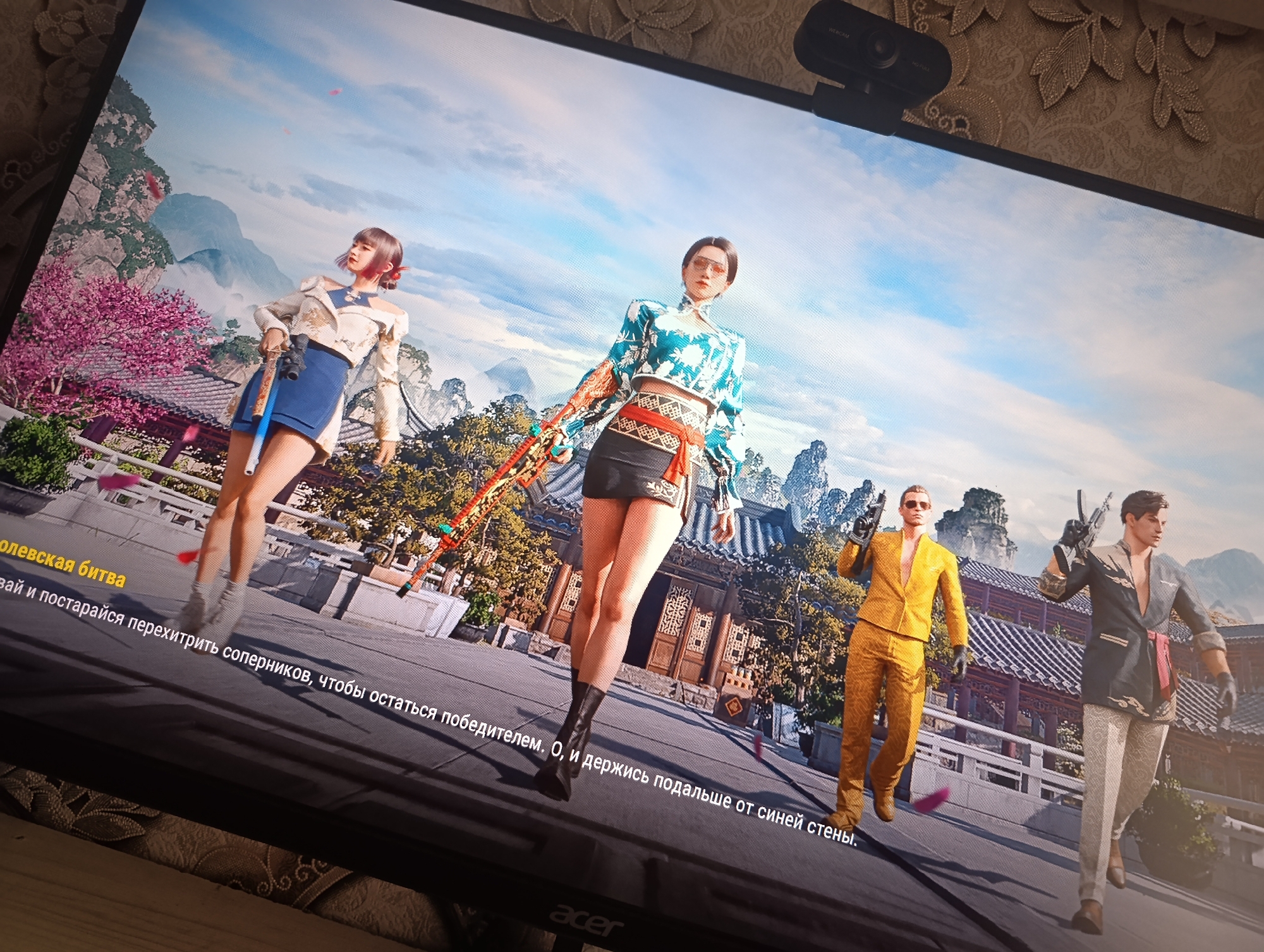 A digital screenshot displays a video game scene. Four characters are visible in the foreground, rendered in a realistic style. On the left, a woman with short dark hair and a red ribbon in her hair wears a white and gold jacket with an off-the-shoulder design, a blue skirt, and brown boots. She is holding a rifle. Next to her, a woman with dark hair pulled back wears a teal and white patterned crop top, a black skirt with a red sash, and black boots. She holds a rifle. Further to the right, a man in a bright yellow suit, sunglasses, and brown shoes walks while holding a pistol. To his right, a man in a dark grey jacket with intricate patterns, white pants, and brown shoes holds a pistol. The background depicts a stylized Asian-inspired environment with ornate buildings, a bridge, and mountainous scenery under a blue sky with clouds. The overall scene suggests an action or combat scenario within a game. There is no discernible connection to Dushanbe, Tajikistan.