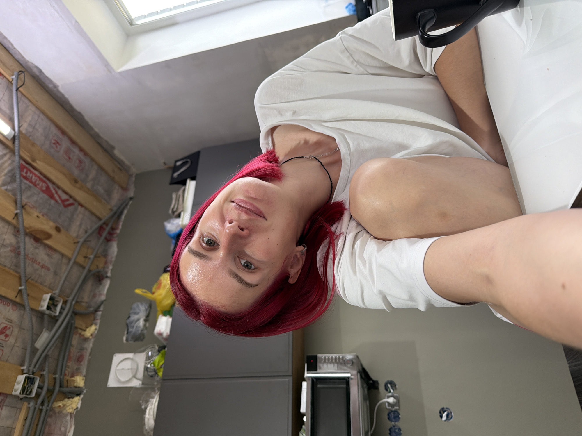 A selfie-style photograph captures a young adult with vibrant red hair and fair skin, looking directly at the camera with a gentle smile. They are wearing a white t-shirt and a delicate black beaded necklace, and a black mug is partially visible near their head.
The setting appears to be an indoor space undergoing renovation, likely a kitchen or utility room in Chaikovsky, Russia. Natural light streams in from a skylight or large window in the ceiling, which appears to be freshly plastered or unfinished.
On the left side, a wall is partially stripped, revealing wooden studs and insulation material with visible text including "SMART" and "utia.pro". Exposed electrical conduits and junction boxes are fastened to this wall. On the right side, modern dark grey cabinet units are installed. A metallic appliance, possibly a toaster oven or microwave, sits on a countertop. Below the countertop, electrical outlets are visible, one with a white cable plugged in, and an empty circular cutout for another outlet. The overall impression is a room in transition, blending new installations with ongoing construction. The time of day is clearly daytime due to the bright natural light.