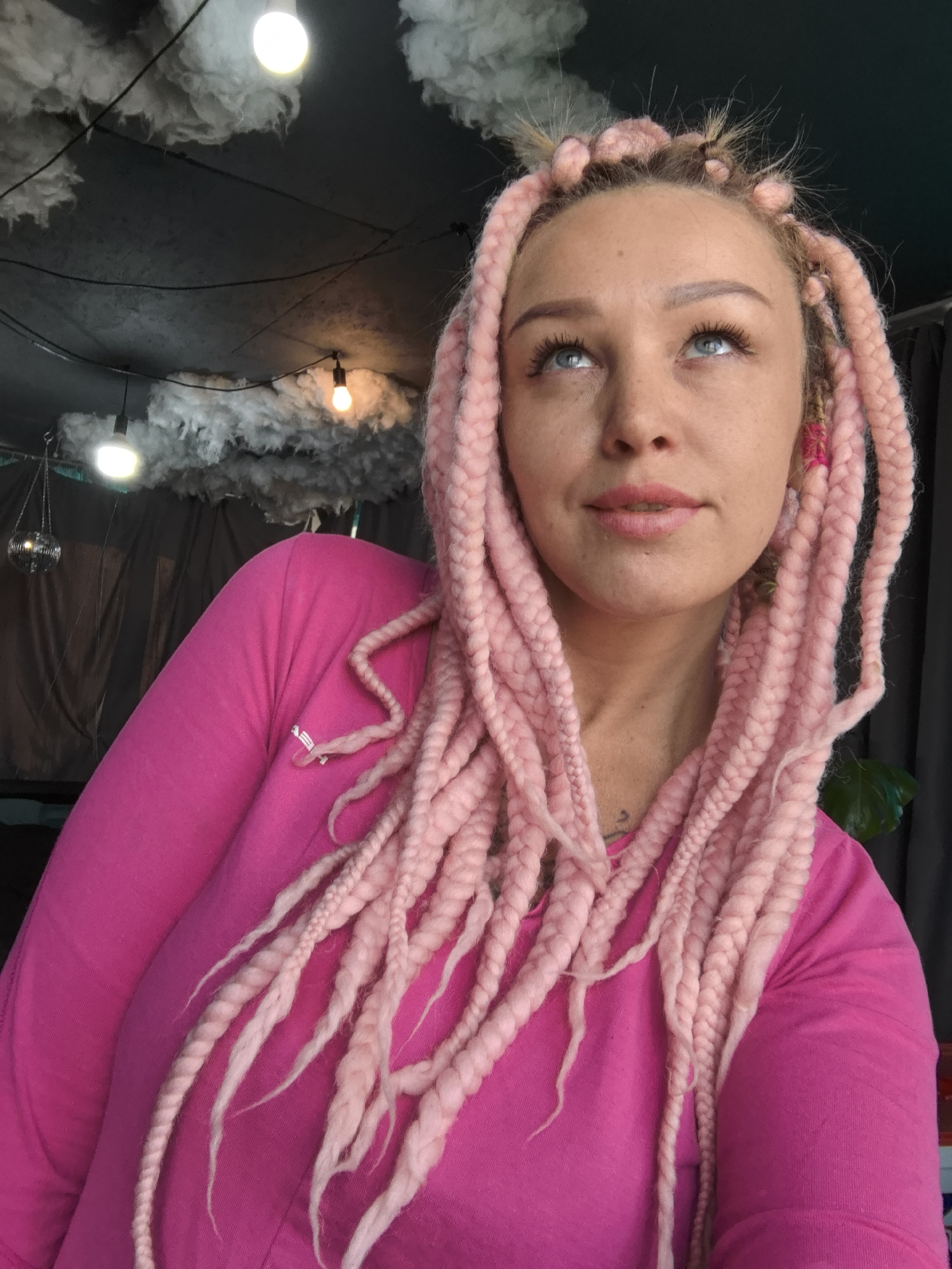 A woman with pink braids looks upward in a room decorated with artificial clouds and hanging lights. The room's ceiling is partially visible, showing a dark surface adorned with the cloud-like decorations and illuminated by bulbs. A disco ball hangs in the background to the left. The woman wears a pink long-sleeved shirt. Her eyes are blue, and her expression is thoughtful. Part of a green leafy plant is visible on the right side. There is no indication that the location is Noviy, Russia.