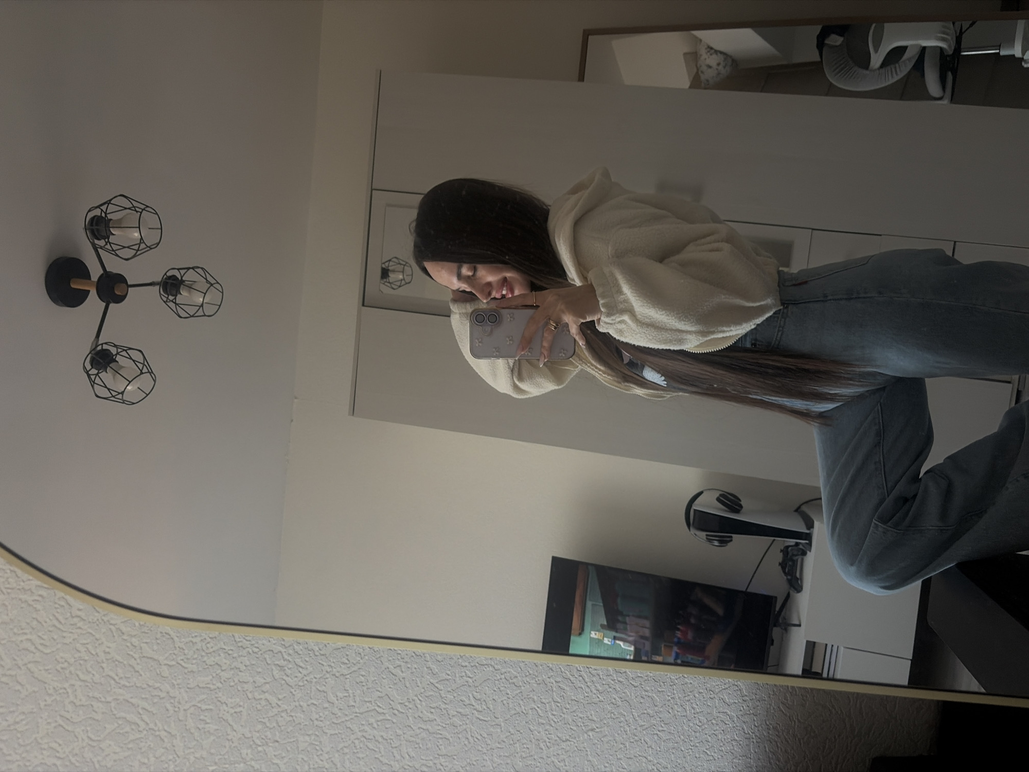 A young woman with long brown hair and wearing a white fleece jacket and blue jeans is taking a picture of herself in a mirror. The reflection shows a living space with a TV, a video game console (likely a PlayStation 5), and headphones on a shelf. A modern black chandelier with caged bulbs is mounted on the wall near the mirror. The room appears to be well-lit and simply decorated. There's no information available from the image to infer a location in Saint Petersburg.