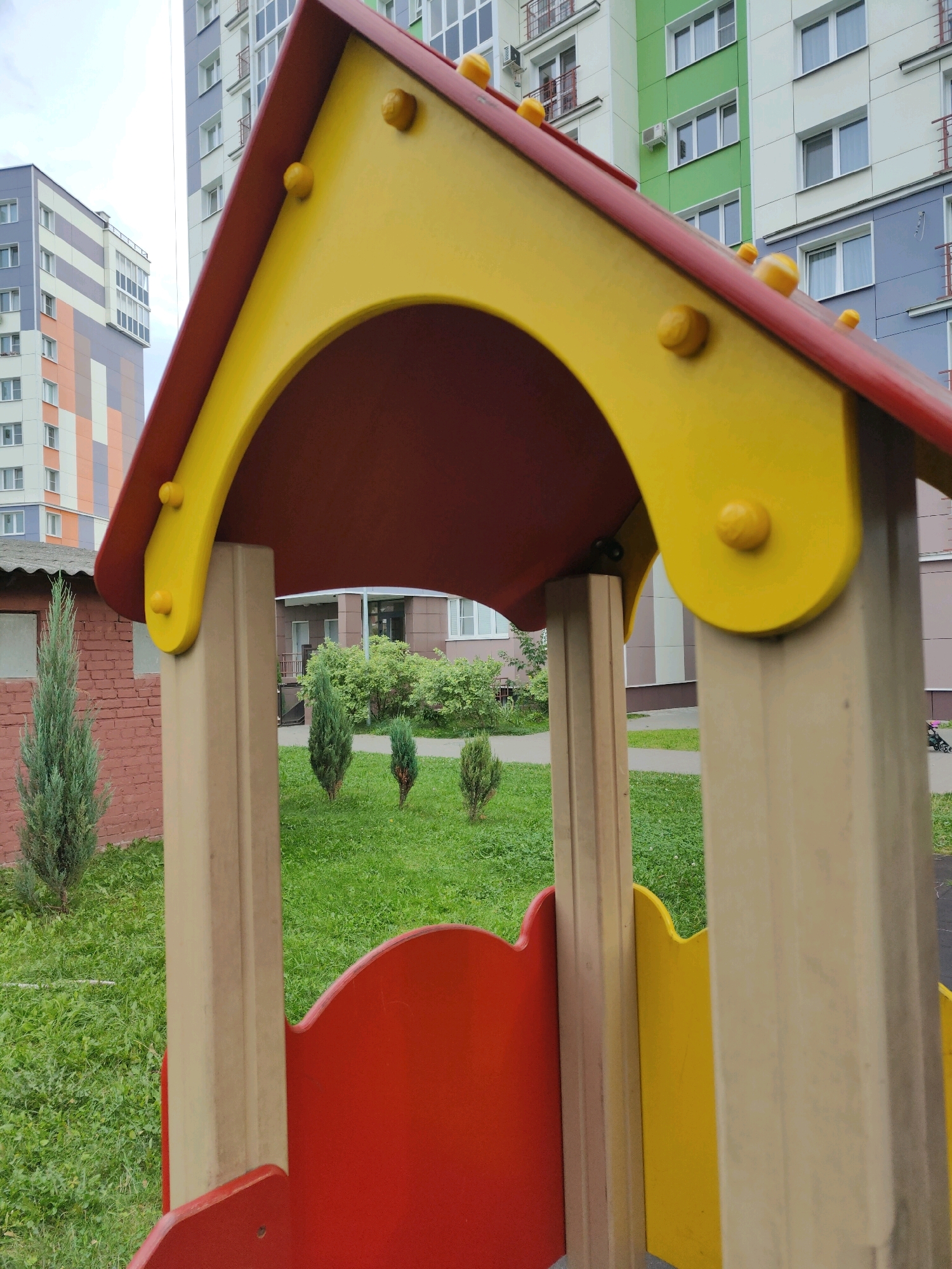 In the city of Homyel, Belarus, a close-up, eye-level shot features a colorful, wooden children's playhouse with a yellow, arched roof and red and yellow panels. The playhouse is situated in a grassy area with several small evergreen trees. In the background, modern apartment buildings with various colored facades and numerous windows are visible, suggesting a residential urban environment. The sky is overcast. There are no people or animals visible in the image.