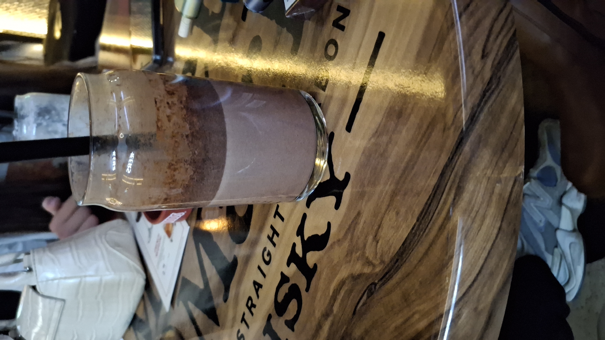 The image shows a chocolate milkshake in a glass with a black straw, sitting on a wooden table with the word "STRAIGHT WHISKY" printed on it. Beside the glass is a white purse, a menu, and a pair of sneakers. The location is Unknown, Unknown.