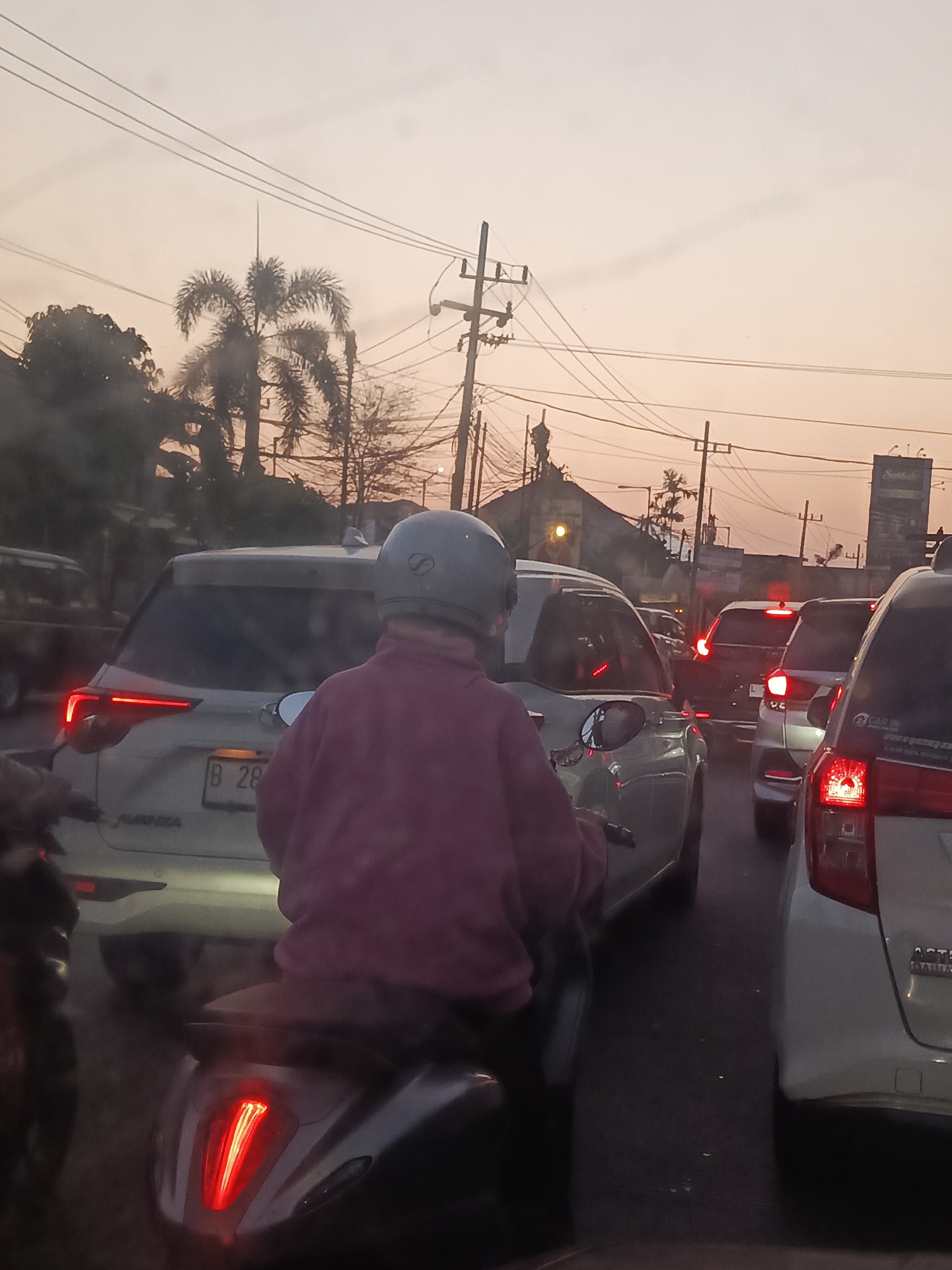 The image shows a traffic jam on a street in Sidoarjo, Indonesia, during sunset. A person on a motorcycle, wearing a grey helmet and a pink jacket, is in the foreground, facing away from the camera and looking at the traffic. The motorcycle's taillight is illuminated. Several cars are also visible in the traffic jam, including a white SUV with its brake lights on, and other vehicles of various colors. Power lines and utility poles are present in the background, along with trees and buildings. The sky is a pale orange, indicating the setting sun. The overall scene depicts a common urban traffic situation during the evening commute.