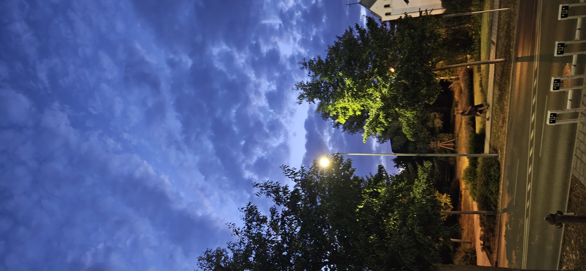 The image shows a street scene in Hévíz, Hungary, at dusk. A street lamp illuminates two trees, one on the left and one on the right. The tree on the right is particularly well-lit, with its leaves glowing green. In the background, a white building with a cross on top, possibly a church, is visible. A person is walking on the sidewalk in front of the well-lit tree. The sky is a deep blue with scattered clouds, indicating the transition from day to night. The street in the foreground has white lines indicating traffic lanes and a fire hydrant is visible on the left side of the road. Several metal stands are positioned along the sidewalk in the lower right portion of the image.