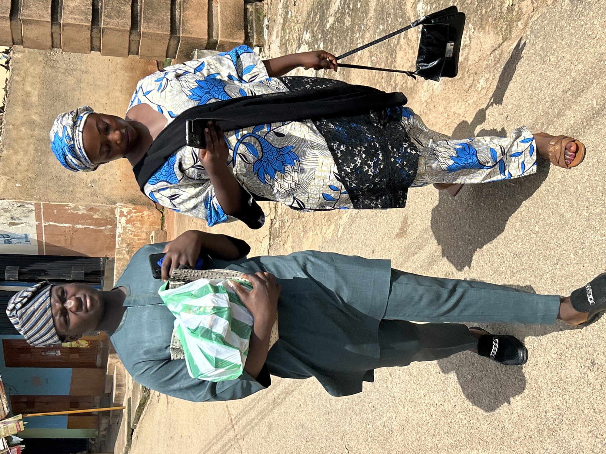 In Jos, Nigeria, a man and a woman walk on a paved street under a bright sun, casting distinct shadows. The man, on the left, wears a teal, traditional African outfit with a matching cap adorned with blue and white stripes. He carries a light-colored basket filled with items, one of which appears to be a green and white striped bag. He holds a black smartphone in his right hand and wears black slip-on shoes with a silver chain detail. The woman, on the right, wears a blue and white patterned traditional African dress with a black sash draped over her shoulder. She also wears a matching headscarf. Her dress features a black lace trim along the hem. She holds a black smartphone in her left hand and carries a black, rectangular handbag with a silver clasp on her right shoulder. Her right foot is adorned with a brown sandal, and her left foot is not clearly visible. The background features a rustic building with brown walls and elements of a street, including a weathered wall and a doorway.