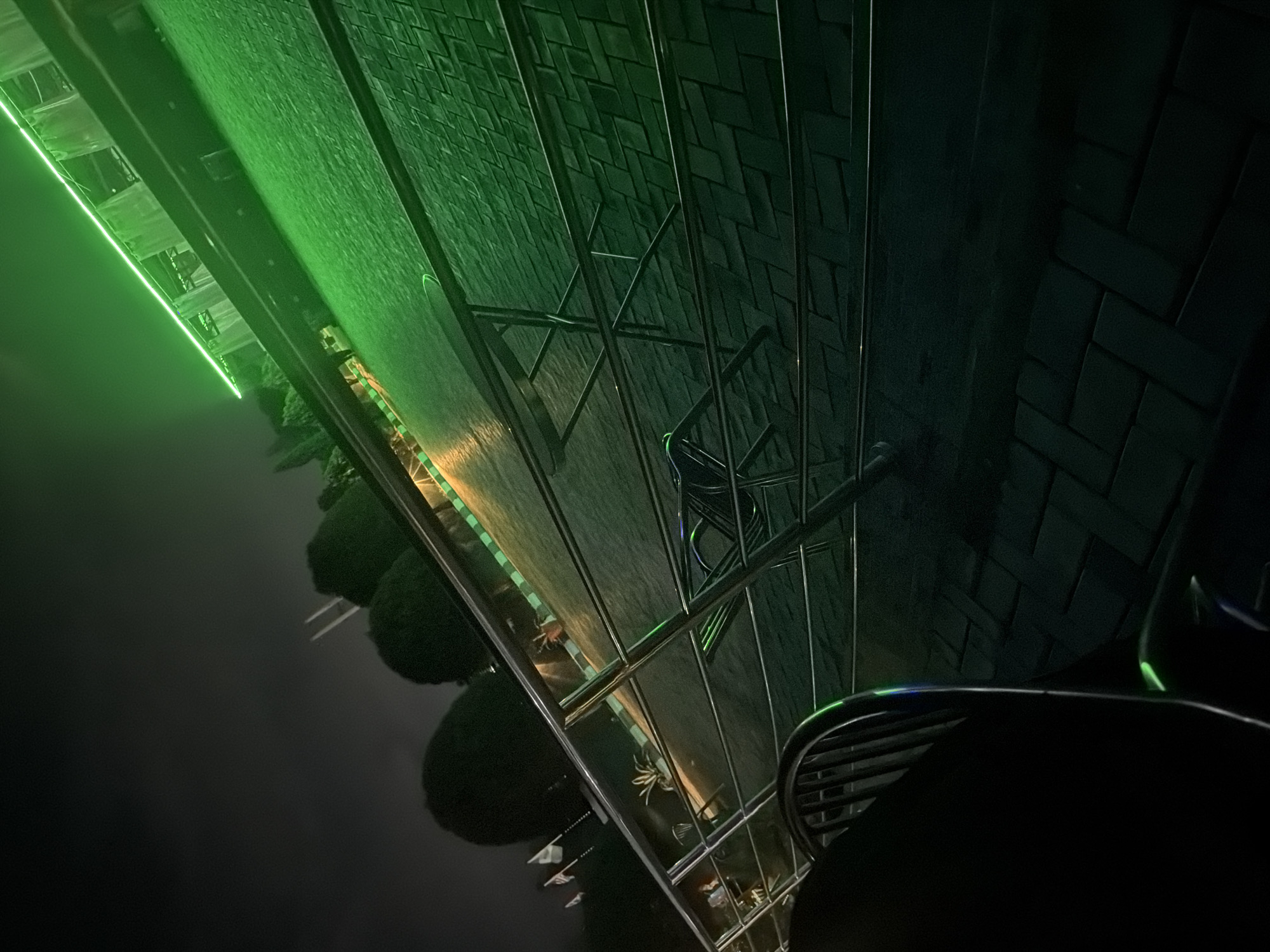 The image depicts an outdoor scene at night, likely in Bukuru, Nigeria, characterized by ambient green lighting. In the foreground, a dark, rounded object, possibly a table, occupies the lower left corner, with a black chair with vertical slats visible to its right. A metallic railing, composed of horizontal and vertical bars, stretches diagonally across the mid-ground. Beyond the railing, a paved surface is illuminated by the vibrant green neon lights that line the upper edge of the frame, casting a strong glow on the textured pavement. In the background, silhouetted trees with rounded canopies are visible against the dark sky, along with several flags on poles. The overall impression is a nocturnal setting that combines natural elements with artificial lighting, suggesting a public or commercial space.
