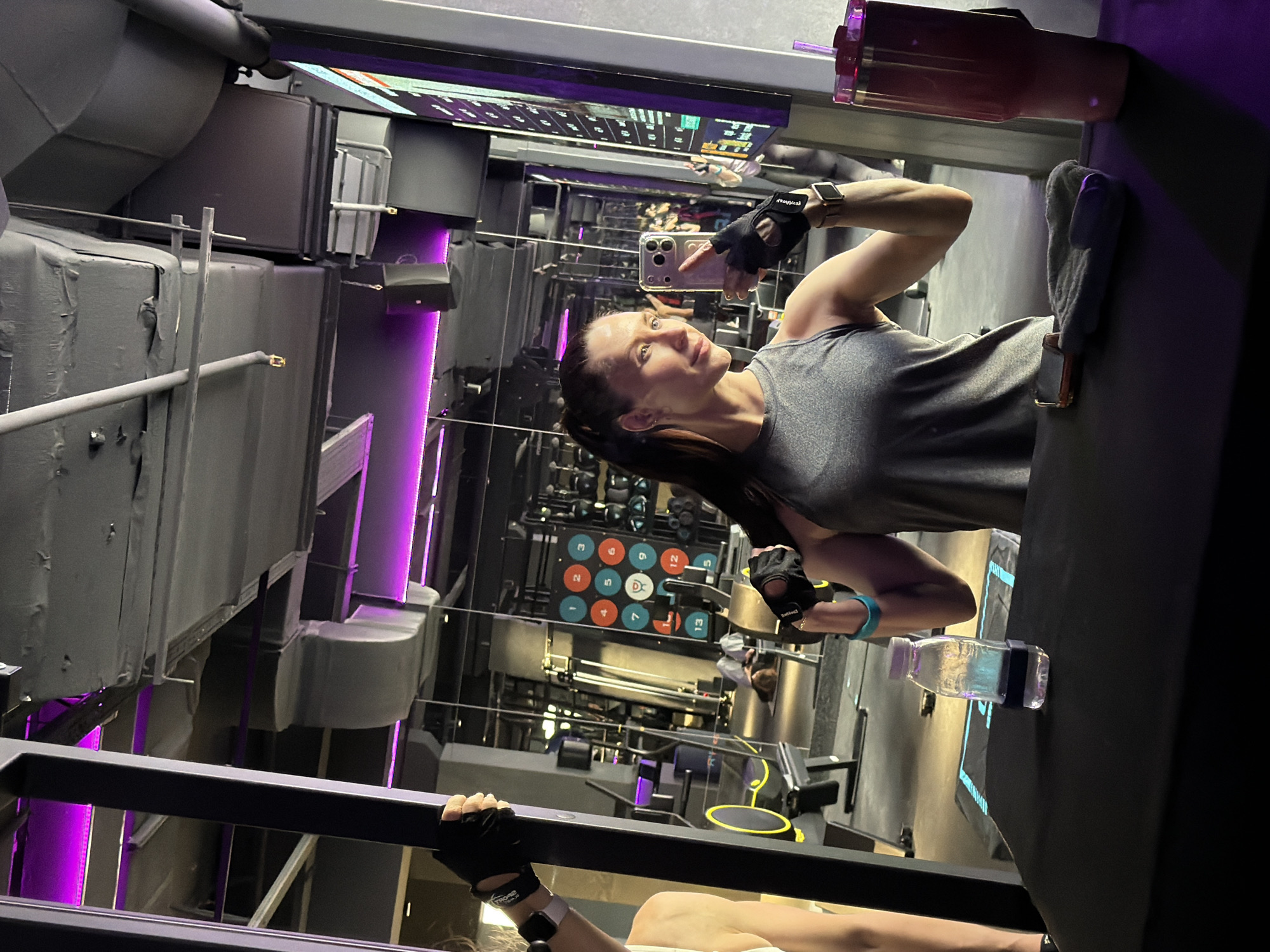 The image captures a woman in a modern gym located in Perm, Russia, seemingly taking a selfie or video during a workout. She is seen from the chest up, with her dark hair pulled back in a ponytail, looking upwards and slightly to her right with a focused expression. She wears a grey athletic tank top, black fingerless gym gloves (one bearing partially visible text like "BODYTRAIN"), and a smartwatch on her left wrist. Her right hand holds a light-colored smartphone with a multi-camera setup.
She appears to be positioned on a piece of gym equipment, possibly a bench, with her left hand holding onto a black horizontal bar in the foreground. Nearby, on the equipment surface, are a clear plastic water bottle with a light purple cap, a grey towel, and another smartphone. A large, reddish-pink opaque drink bottle with a straw is also visible towards the top right.
The gym environment features an industrial aesthetic, with dark grey exposed ductwork and ceiling beams, dramatically lit by vibrant purple LED strip lights. The background is dominated by reflective surfaces, likely mirrors, which reflect other gym equipment. Visible in the reflections are colorful circular targets with numbers (e.g., 1, 2, 5, 9, 12, 13) and letters (C, D) on a wall, along with other exercise machines. The lighting is artificial and creates a moody atmosphere, suggesting it might be evening or night. The image is taken at an angle, as if the camera was held sideways.