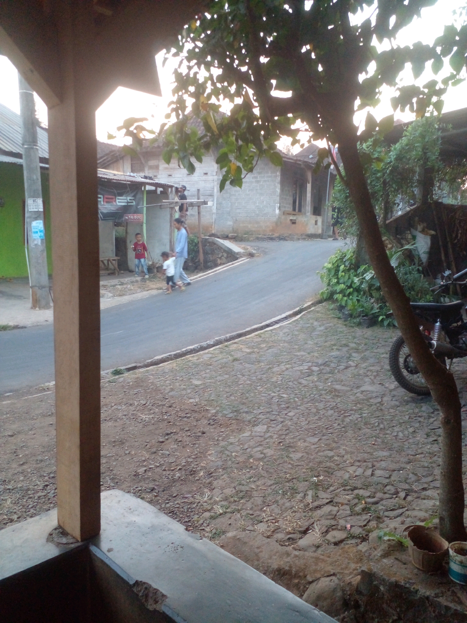 The image depicts a street scene in Pungangan, Indonesia, with a focus on a paved road and adjacent areas. In the foreground, a wooden pillar and a concrete structure are visible, suggesting the presence of a building or porch. Across the street, a group of people, including two children and an adult woman, are walking. One child is wearing a red shirt, and the other is in white. The adult is in a light blue outfit. Behind them, a small building with a dark awning and a sign with text "ARES" is partially visible. Further down the street, more buildings constructed with cinder blocks line the road. To the right of the scene, a dirt and gravel area is present, featuring a tree and a parked motorcycle with visible suspension. The ground is covered with stones and some vegetation. The overall lighting suggests it is either early morning or late afternoon.
