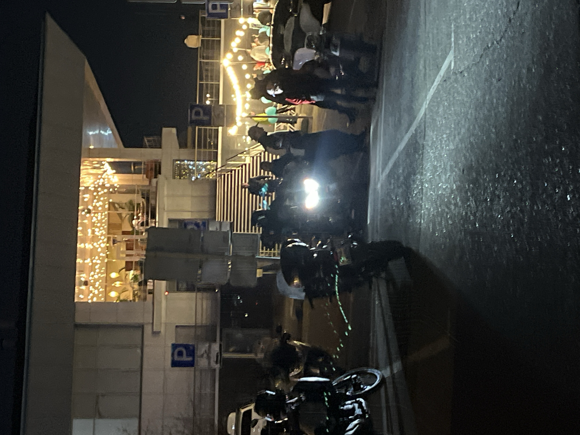 The image captures an outdoor night scene beside a modern building, likely a commercial establishment or leisure venue. In the foreground and midground, a group of people are gathered around several motorcycles and at least one bicycle. One motorcycle features a prominent, bright headlight shining forward. The individuals are mostly obscured by darkness and distance, but appear to be standing and interacting, suggesting a social gathering or meet-up.
To the left, the multi-story building has large windows and a balcony or terrace on an upper level, which is brightly illuminated with numerous string lights, creating a warm, festive glow. Other parts of the building also show interior lights. Several blue signs with a white letter "P" are visible on the building's facade and railings, indicating parking areas. The ground is dark, appearing to be asphalt with some faint markings and texture, possibly reflective or slightly damp. The overall atmosphere suggests a lively night event or social occasion.