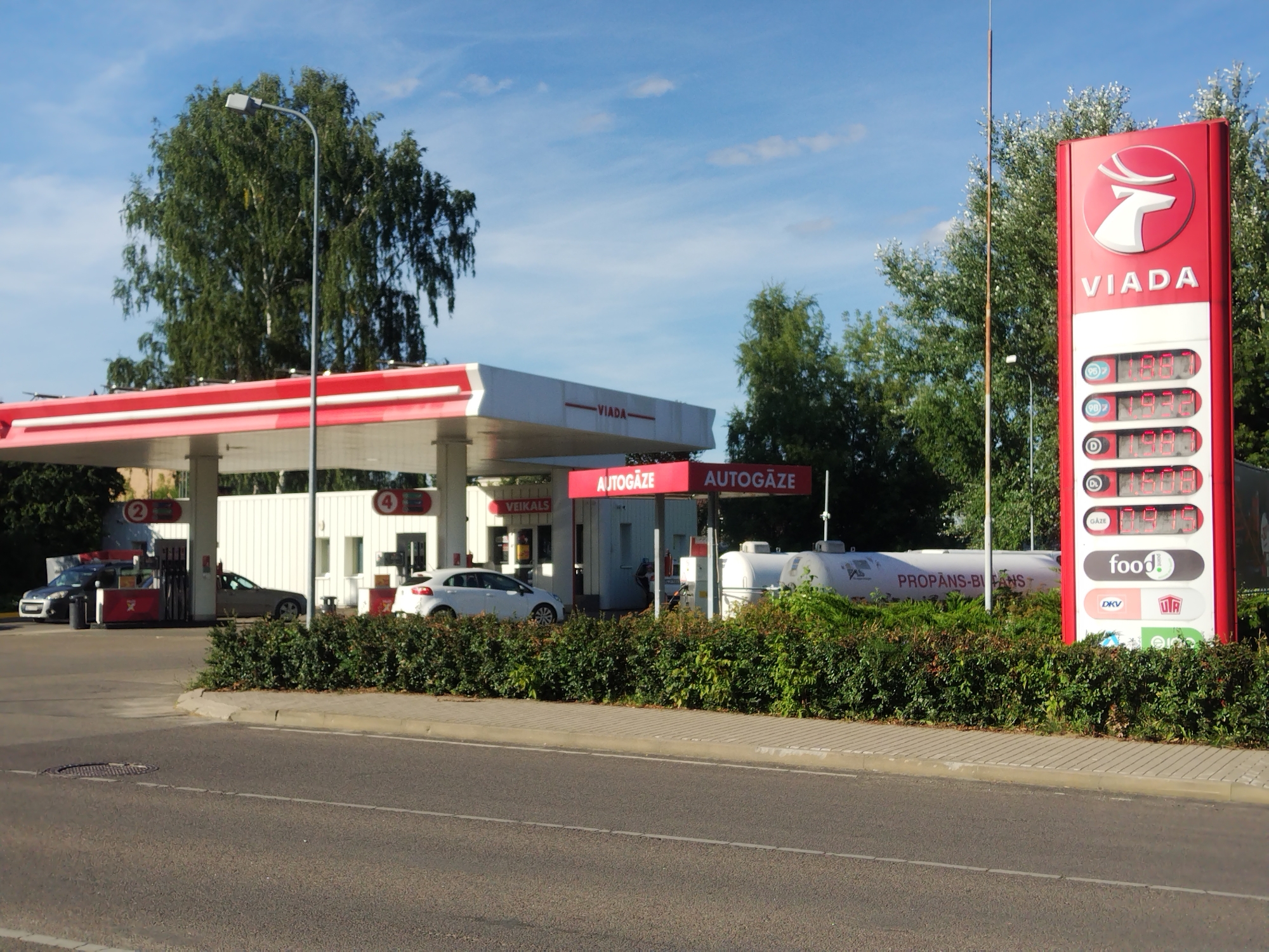 This is a daytime exterior, eye-level, medium shot of a Viada gas station. The scene is set on a street in Daugavpils, Latvia. The foreground shows a paved road with a light gray sidewalk and a curb. Beyond the sidewalk, a dense, green hedge lines the front of the gas station.
The gas station structure is white with red trim. A red and white pylon sign for Viada stands prominently on the right side of the image, displaying fuel prices. The prices visible are 1.188 for two different fuel types and 1.198 for a third. Below these are logos for "food!" and other entities. To the left of the pylon, a large white propane tank is visible behind the hedge, with the text "PROPANS-BI-ANS" printed on it.
Underneath the gas station canopy, several vehicles are present. A dark grey car is parked at a pump on the left. Further to the right, a silver car is parked. A white car is positioned near a pump further back. Two red signs with the word "AUTOGĀZE" are mounted on the canopy. A smaller sign that reads "VEIKALS" is also visible. The background consists of trees and a blue sky with some clouds. A lamppost stands to the left of the Viada sign.