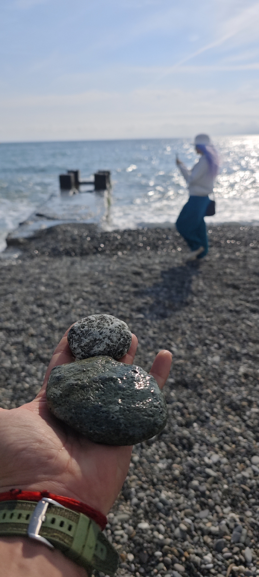 The image depicts a hand holding two smooth, wet rocks on a pebble beach. The hand is adorned with a red string bracelet and what appears to be a wristwatch. The background features a blurred figure, likely a woman, wearing a white hat, light-colored sweatshirt, and teal pants, seemingly taking a picture with a phone. Behind her, the sea glistens in the sunlight, with a partially submerged concrete structure in the water. The beach consists of small, grey pebbles, typical of the Black Sea coastline. Based on the environment, the location is likely Sochi, Russia.