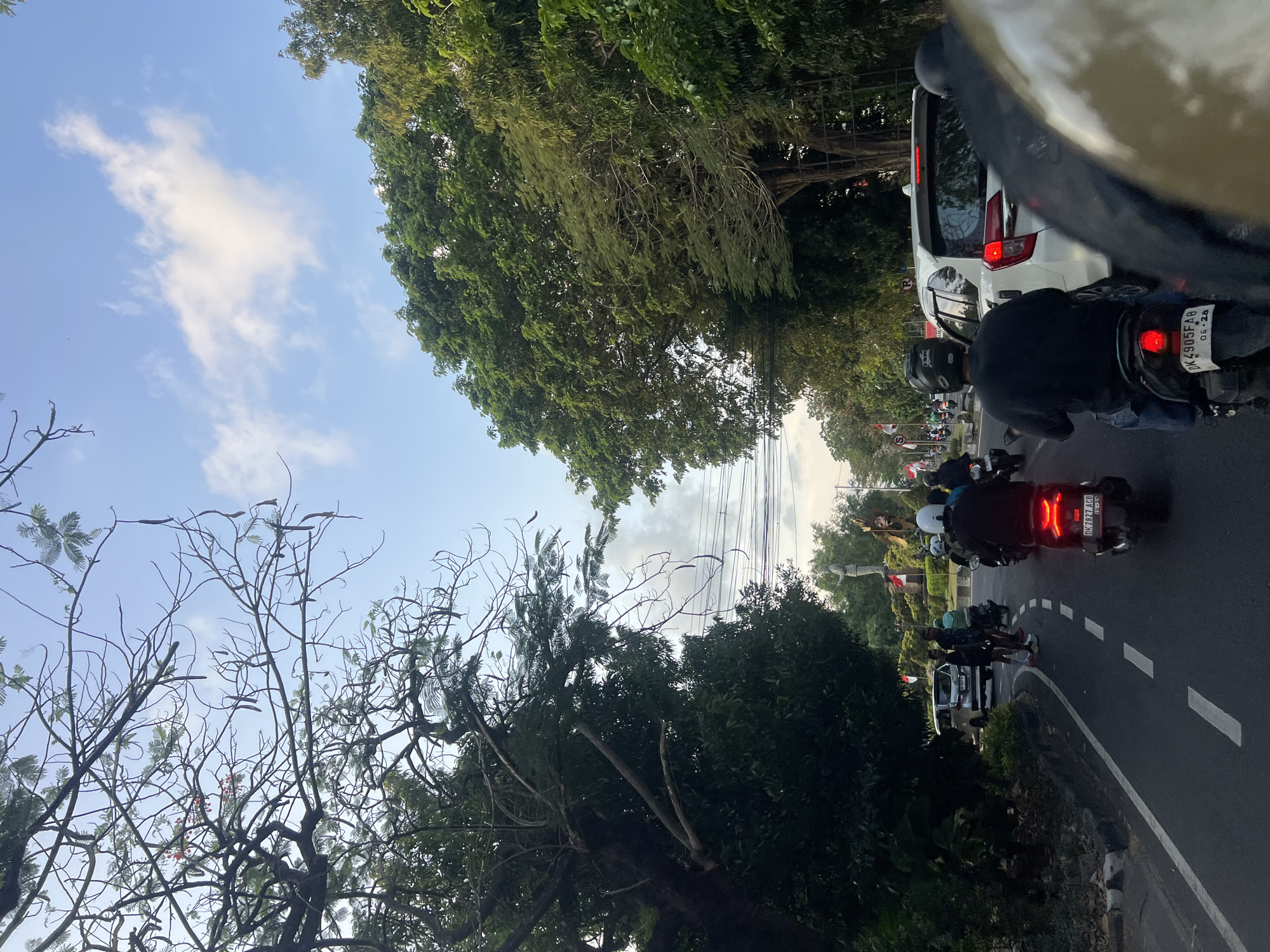 The image depicts a street scene in Denpasar, Indonesia, characterized by traffic congestion and lush greenery. In the foreground, several motorcycles are stopped or moving slowly in traffic. A person wearing a black helmet and shirt is riding a black scooter, visible from behind. Another scooter with a red taillight is in front. To the right, a white car is also in traffic, with its brake lights illuminated.
The street itself is paved with asphalt, marked with white dashed lines indicating lanes. On the left side of the road, a large tree with dense foliage overhangs the street, its branches reaching towards the sky. Further down the street, a white van is visible.
In the background, there is a statue or monument, and several Indonesian flags are displayed, suggesting a public space or event. The sky is partly cloudy, with a mix of blue sky and white clouds, and the overall lighting suggests it is either early morning or late afternoon. The environment appears tropical, with abundant trees and vegetation lining the road. Power lines are also visible overhead.
