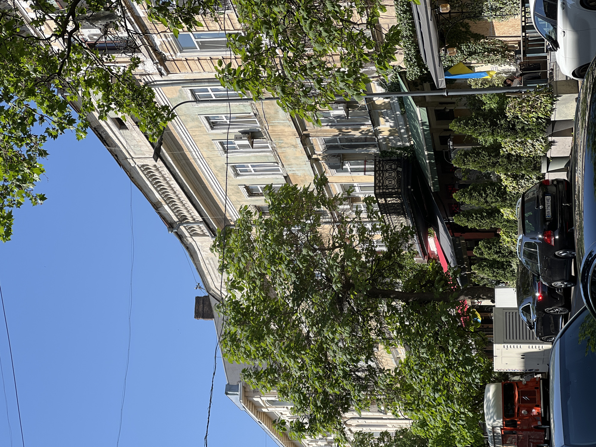 The image captures a street scene in Odesa, Ukraine. A multi-story building, likely residential or commercial, dominates the left side of the frame. Its facade is adorned with windows, architectural details, and a balcony with iron railings. Lush trees partially obscure the building. On the right, a street is lined with parked cars. A red truck is visible. A small blue and yellow flag is hanging from an awning. The sky is clear and blue.
