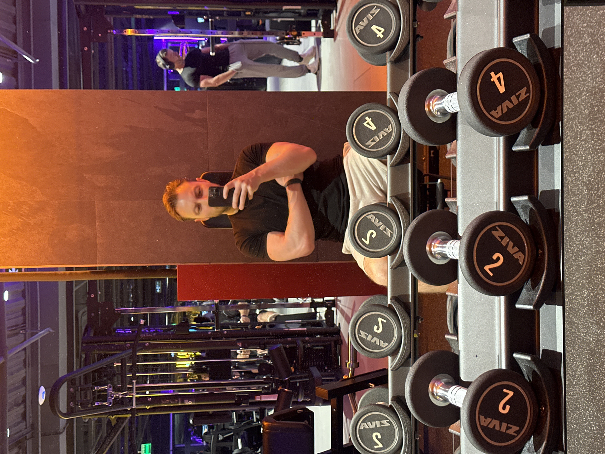 A man in a black t-shirt and beige shorts is taking a selfie in a gym. He is positioned in front of a mirror, which reflects another person exercising in the background. To his right, a rack of dumbbells, labeled "AVIS" and "ZIVA," is visible. The weights range from 2 to 4, arranged in ascending order. The gym features various exercise machines and equipment. Based on the "AVIS" dumbbells, the location is likely in Sochi, Russia.