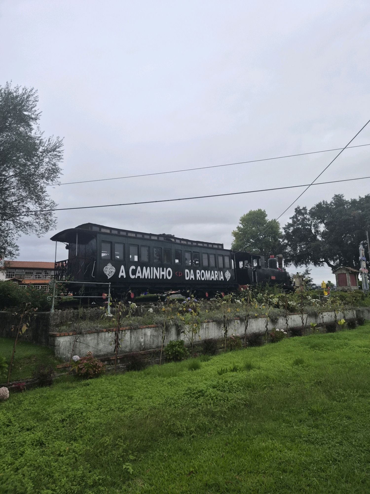 An exterior, low-angle, wide shot captures a vintage, dark green train and carriages adorned with white text reading "A CAMINHO DA ROMARIA" on the side. The train is stationary, positioned on a raised platform. In the foreground, a vibrant green lawn slopes gently downwards, bordered by a low, concrete wall with a brick trim. Behind the wall, a garden of various plants and flowers, some with yellow petals, is visible. A mature tree with dark green foliage stands to the left of the train, and larger trees with similar foliage are situated behind it. A building with a reddish-tiled roof and white walls is visible in the background to the left. The sky is overcast and grey. The scene appears to be located in Darque, Portugal, as indicated by the text on the train.