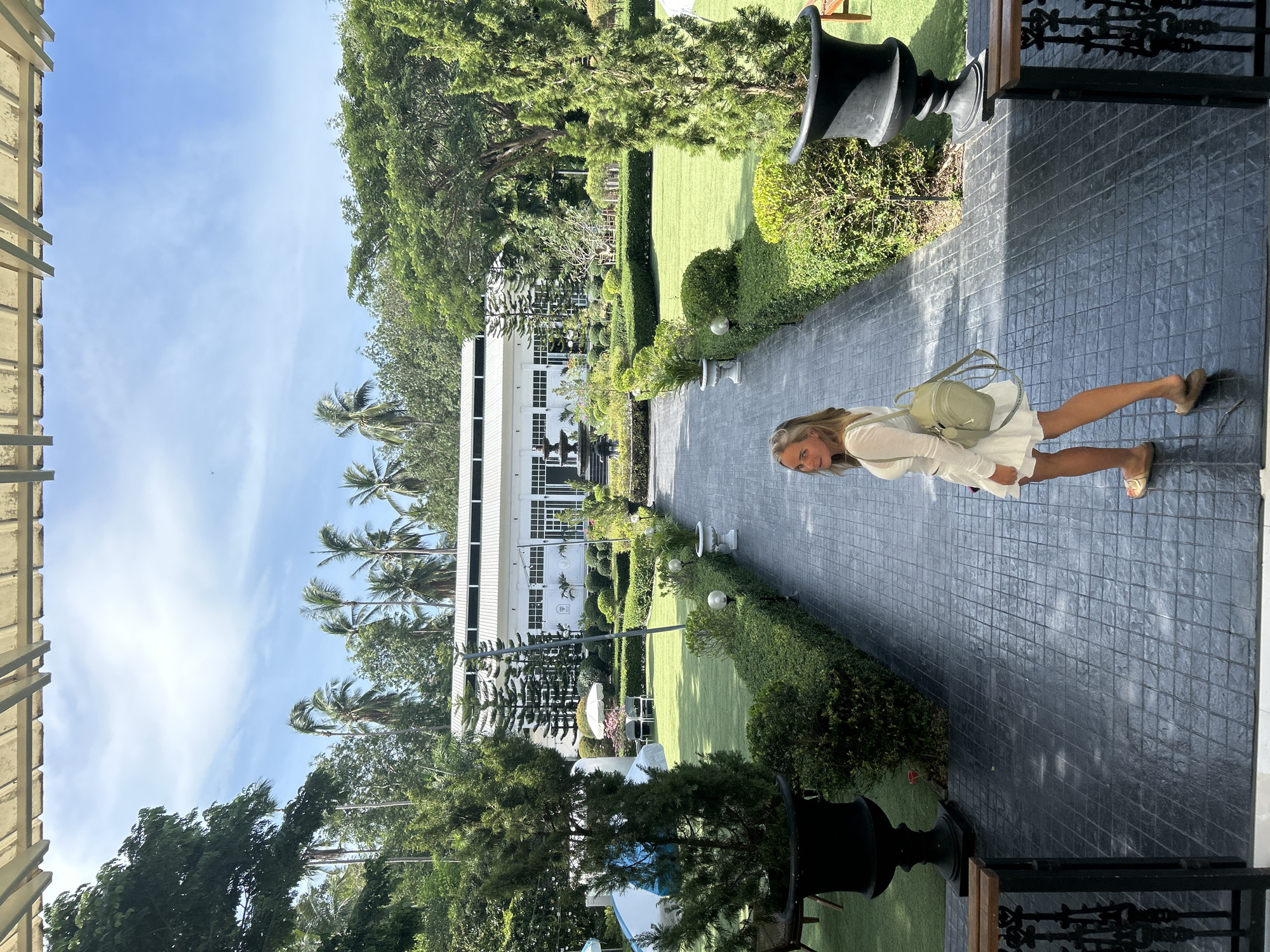 A blonde woman in a white dress and a light green backpack walks away from the camera on a tiled pathway. She is looking over her left shoulder towards the camera. The pathway leads to a fountain and a large white building with a white roof. The building is surrounded by lush green lawns, manicured hedges, and mature trees, including palm trees. There are large black planters on either side of the pathway. The scene appears to be a well-maintained garden or resort area in a tropical location. The sky is blue with scattered clouds.