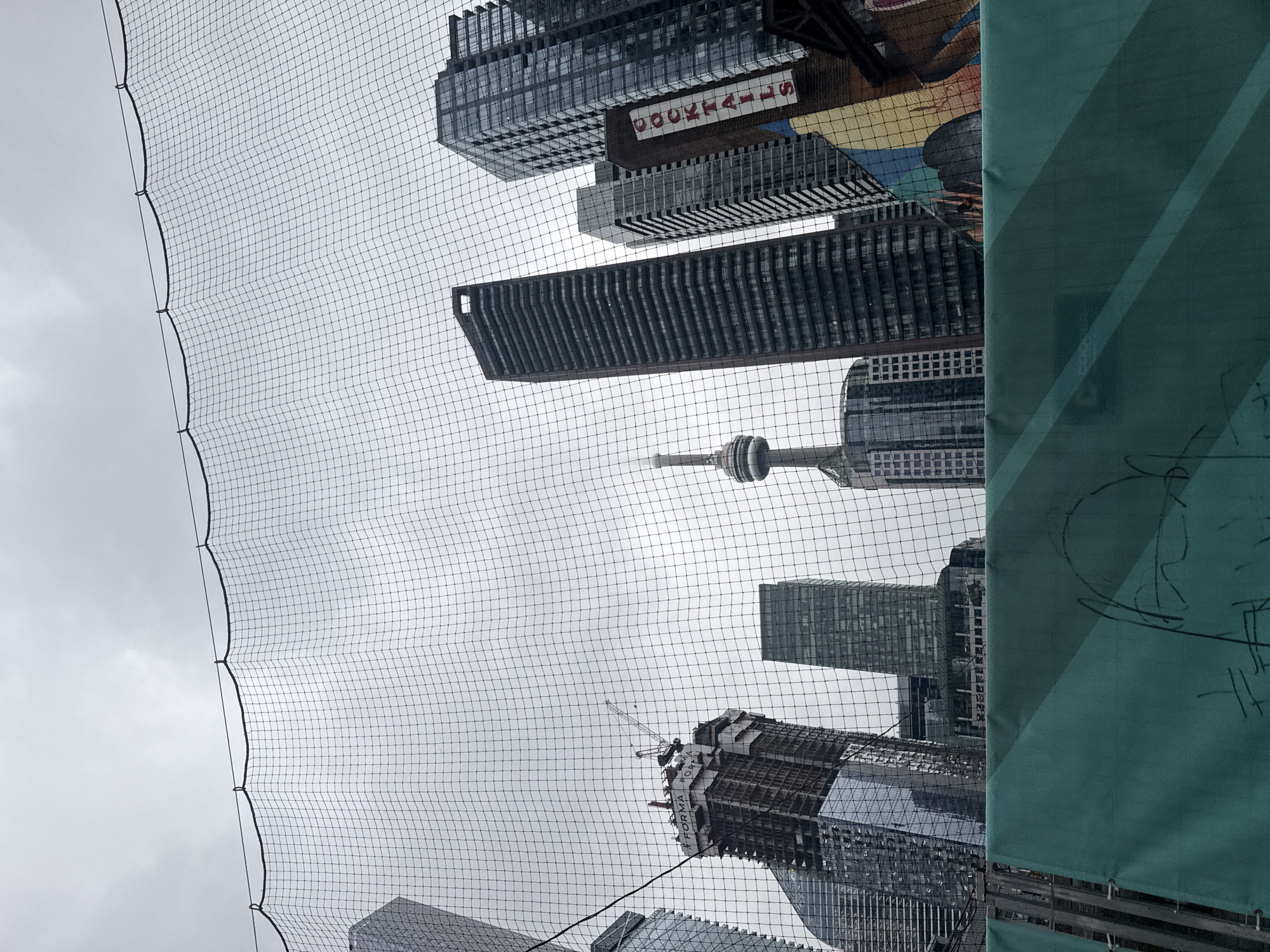 This is a ground-level, wide-angle photograph taken in Toronto, Canada, looking up at a cityscape dominated by skyscrapers. The foreground is partially obscured by a turquoise construction barrier with some faint graffiti. Above the barrier, a black netting material stretches across the frame, with a wire running along its top edge, secured by grommets.
The netting filters the view of the city beyond. Visible in the midground are several tall, modern buildings of glass and steel, some appearing to be under construction. One building in the center-left is clearly a high-rise under active construction, complete with a crane. To the right, the iconic CN Tower is visible, its distinctive silhouette piercing the overcast sky. Further to the right, another cluster of skyscrapers rises, with one building featuring a vertical sign that reads "COCKTAILS" in white letters on a red background. The sky is uniformly gray and cloudy, suggesting an overcast day. The overall impression is one of urban density and ongoing development.