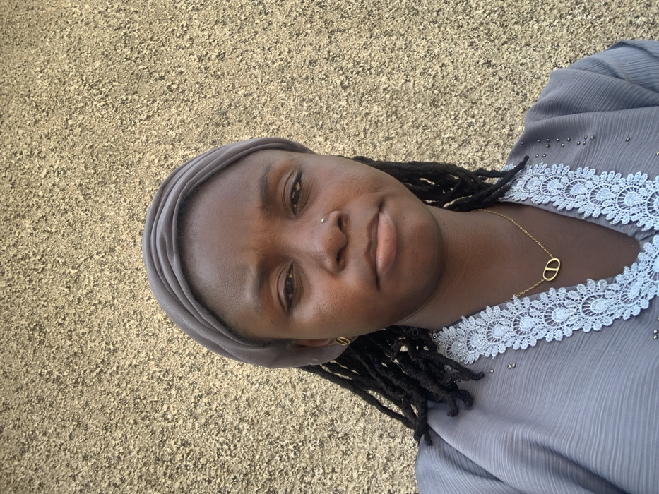 A close-up, chest-up portrait shows a female individual in the city of Bojude, Nigeria, facing the camera directly with a neutral expression. She has dark skin and dreadlocked hair, mostly covered by a grey headwrap. A small silver stud is visible in her left nostril. She wears a grey garment featuring a prominent light blue and white lace trim along the V-neckline. A gold-colored necklace with an intertwined oval 'CD' pendant rests on her chest, and a matching gold-colored open circle earring is visible in her left ear. The background consists of a textured, light brown stucco or rough-cast wall.