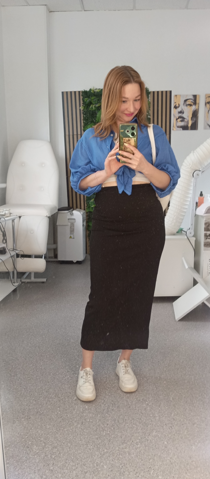 The image depicts a woman in an indoor setting that appears to be a commercial or medical space, potentially a salon or clinic. The woman is fair-skinned with auburn hair, wearing a bright blue, knotted shirt, a black skirt, and white sneakers. She is holding a smartphone with a green case, taking a selfie in front of a large mirror that reflects the room.
Behind her is medical equipment, including a white treatment chair and a white machine. There is a wall with vertical black and tan stripes and artificial greenery. On the adjacent wall, there are framed art pieces depicting faces. The floor is a light gray with a speckled pattern. The location appears to be in a city, but the specific name is not discernible from the image.