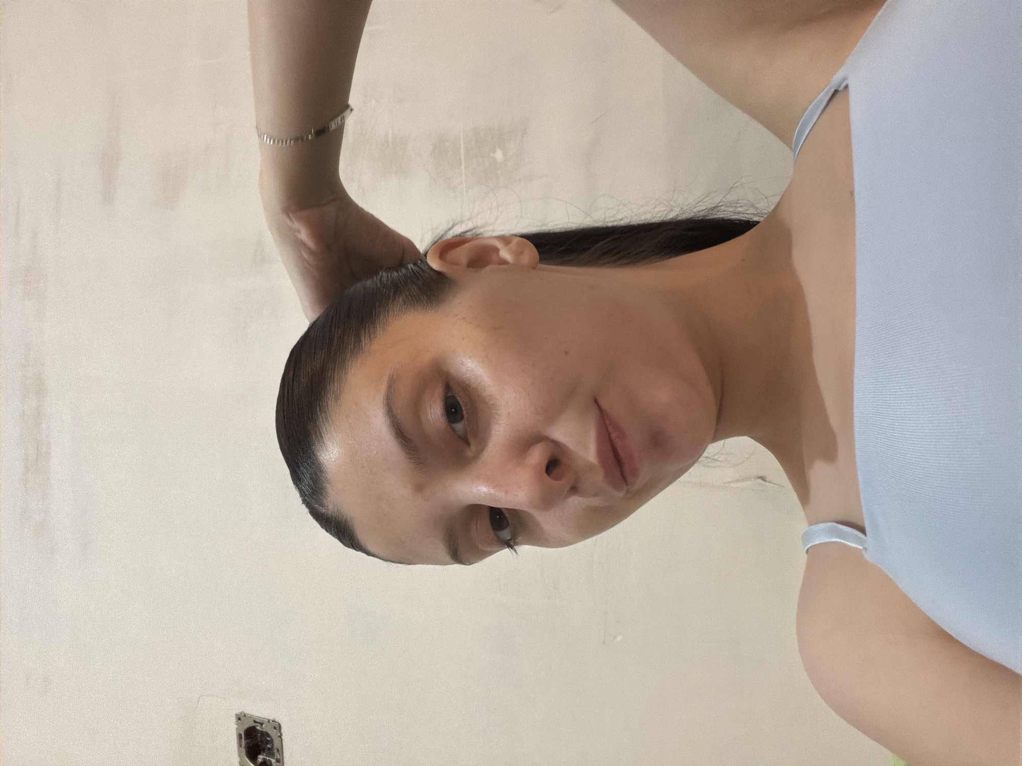 In a close-up, low-angle selfie, a young woman with dark hair pulled back into a ponytail is captured against a neutral, textured wall. She is wearing a light blue ribbed tank top. Her left hand is raised, touching the back of her head, and a thin silver bracelet is visible on her wrist. The background wall appears unfinished, with a visible electrical outlet cover and some minor imperfections. The lighting is soft, highlighting the woman's facial features and the texture of her skin. The scene suggests an indoor, possibly domestic, setting. There is no indication of the specific location within Tbilisi, Georgia.