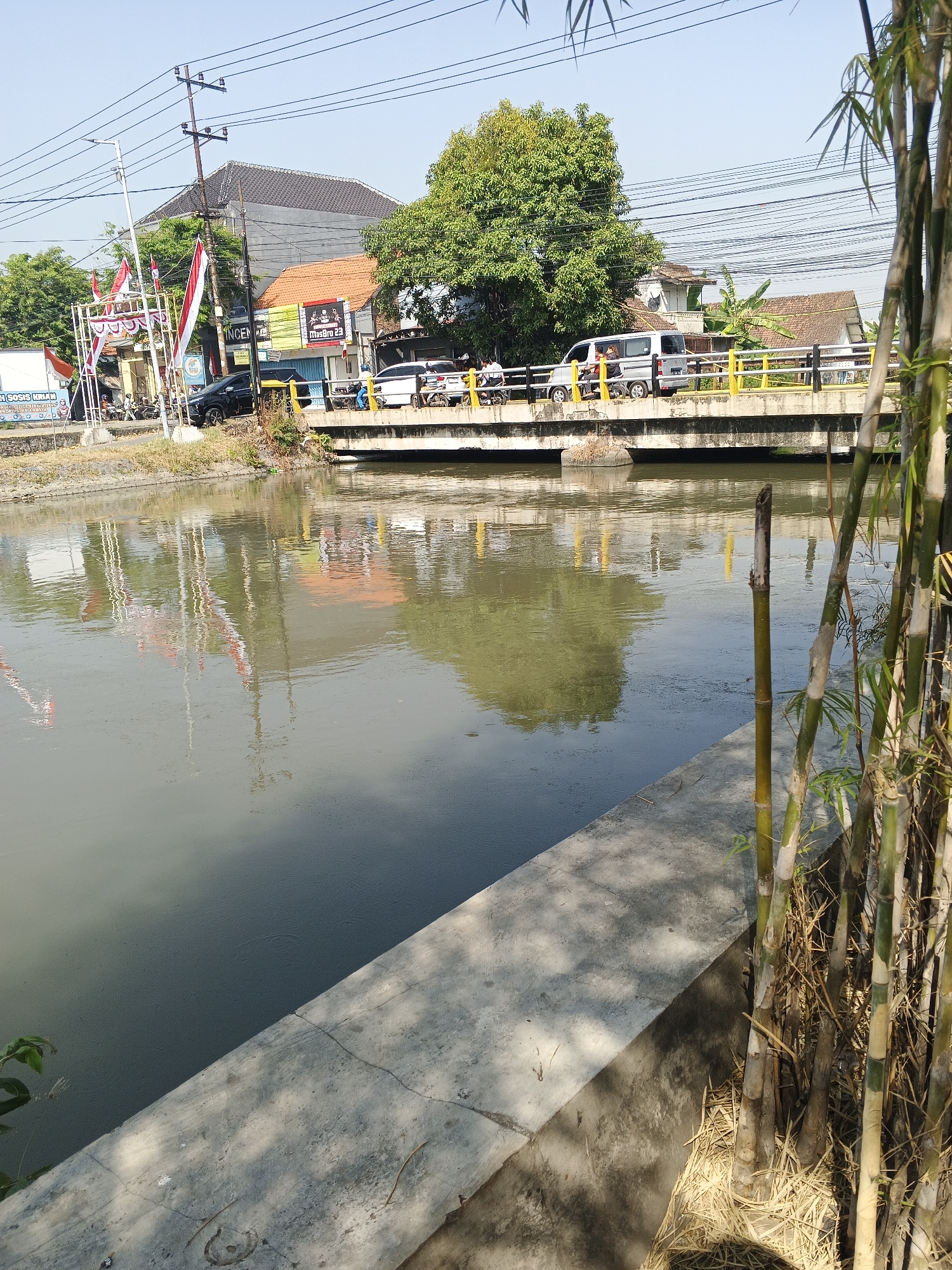 The image depicts a canal in Krian, Indonesia, on a clear, sunny day. A bridge crosses the canal, with several vehicles, including cars and a van, visible on it. Red and white Indonesian flags are displayed on the left side of the bridge. The canal water reflects the surrounding buildings, flags, and trees. On the right bank of the canal, a cluster of bamboo stalks stands tall, with dry grass at their base. The concrete embankment along the canal is visible in the foreground. The sky is bright blue, indicating a cloudless day.
