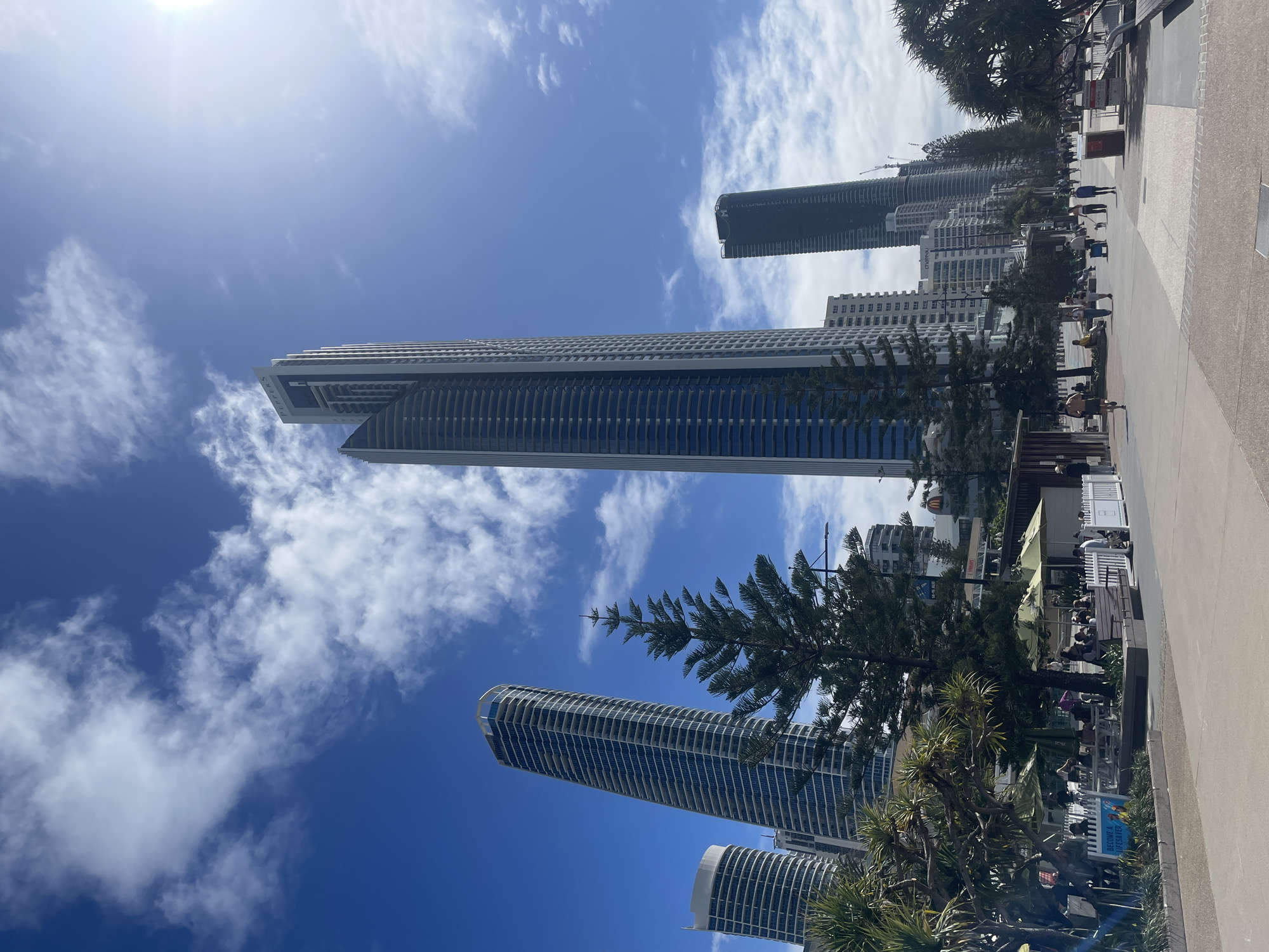 The image shows a sunny day at Surfers Paradise beach in Gold Coast, Australia. The scene features a wide, paved walkway bordering the beach, with a line of palm trees and pine trees providing shade. Several tall, modern high-rise buildings dominate the skyline, including the iconic Peppers Soul Surfers Paradise building.
On the walkway, people are strolling, walking dogs, and sitting at outdoor cafes under green umbrellas. A sign that reads "BECOME A LIFESAVER" is visible in the foreground. The sky is a vibrant blue with scattered white clouds, and the sun is shining brightly. The overall atmosphere is one of a bustling beachside urban environment.