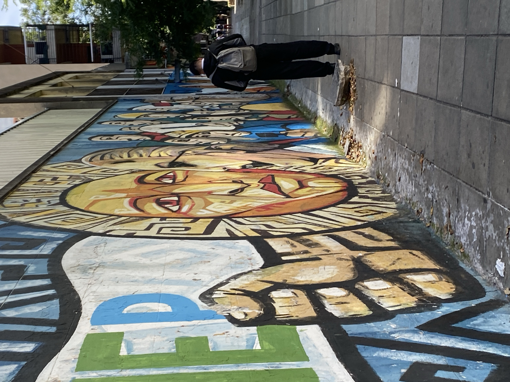 The image shows a mural on a ground-level wall, likely a sidewalk or plaza area. The mural features a stylized depiction of faces and a clenched fist, painted in bold colors including yellow, blue, green, and brown. A person wearing black clothing and a backpack is walking along the sidewalk adjacent to the mural. The location appears to be in Buenos Aires, Argentina, based on the artistic style and common mural art present in the city. The surrounding environment consists of a paved walkway, possibly a street or plaza, and some foliage. There is some evidence of weathering and leaves accumulating along the base of the wall.