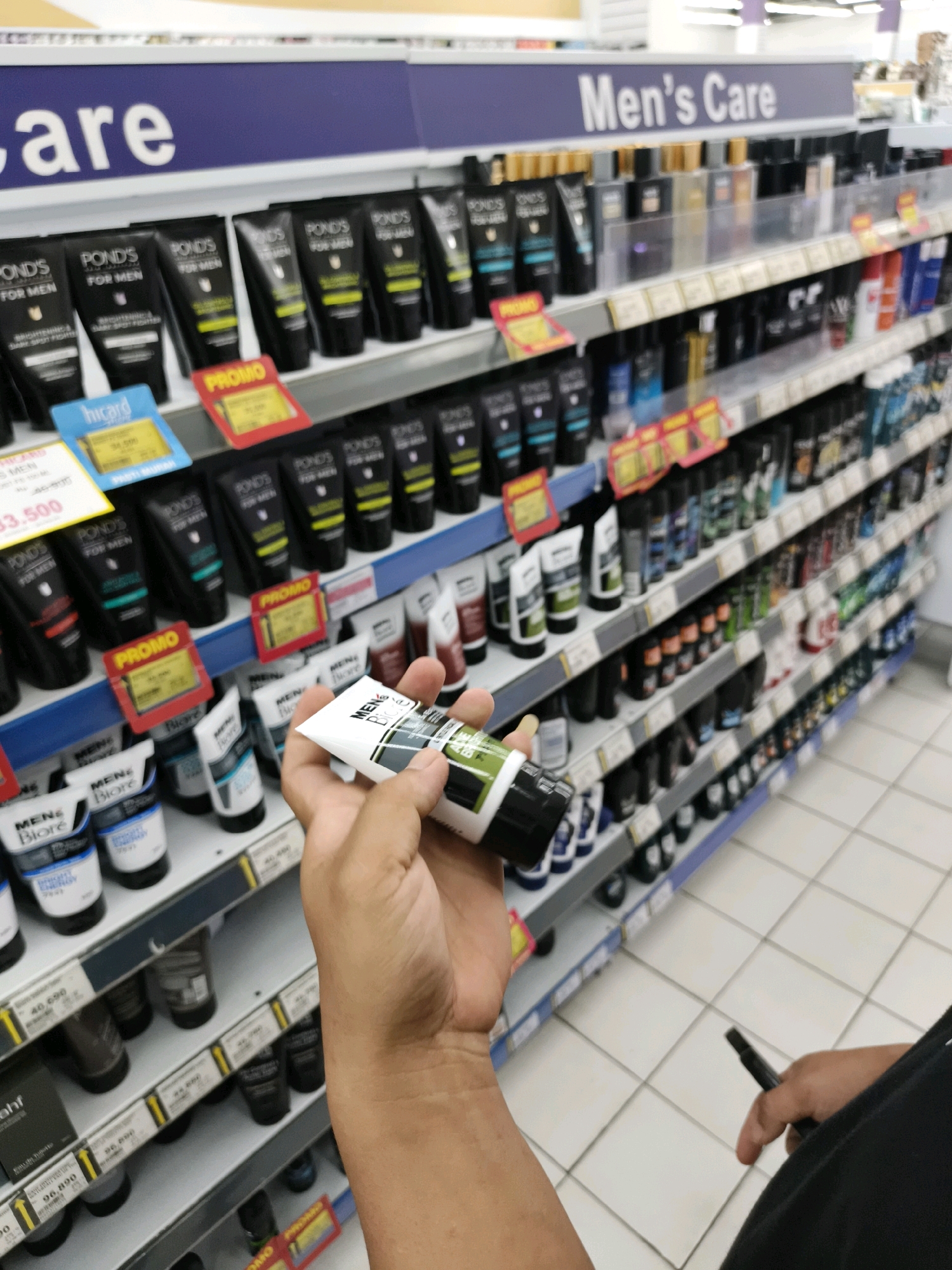 The image shows a person holding a tube of men's skincare product in a retail store aisle. The store appears to be a supermarket or hypermarket, with shelves stocked with various men's grooming products, including lotions, cleansers, and deodorants. The aisle is well-lit, with fluorescent lighting overhead. The shelves are organized by brand and product type. The "Men's Care" sign is prominently displayed above the products, indicating the section of the store. In the background, other shoppers can be seen browsing the aisles. The specific location of the store in Ketapang, Indonesia is not evident from the image, as there are no visible signs or landmarks that would confirm this location.