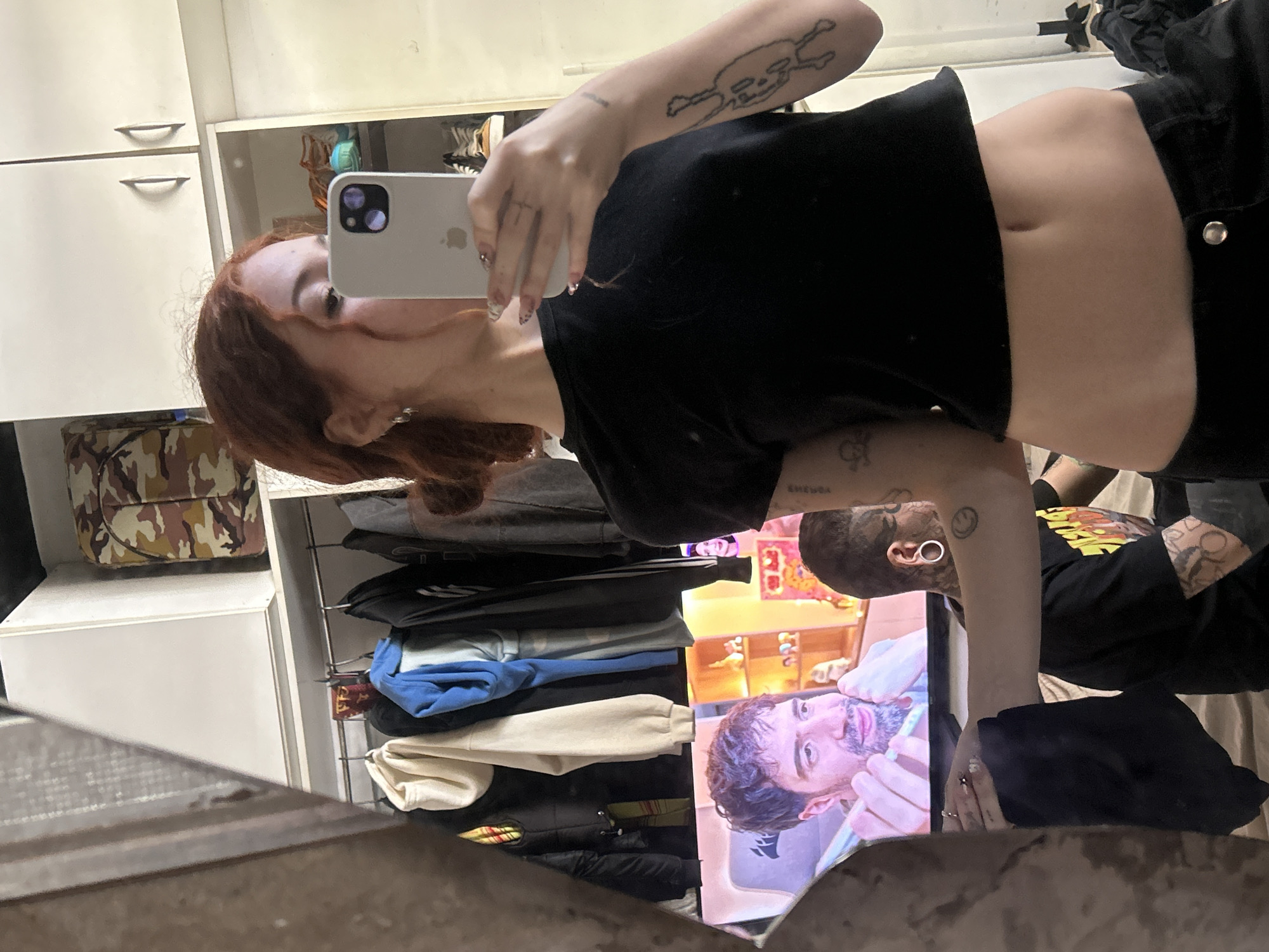 An indoor mirror selfie is presented from a sideways orientation, capturing a person with auburn hair taking the photo. She wears a black cropped top, revealing her midriff, and black bottoms. Her left arm, featuring a skeletal-figure tattoo, holds a light gray iPhone with visible cameras, reflecting her partially obscured face. Her nails are long and manicured.
The background shows white cabinets with silver handles and shelves, on which clothes are neatly stacked, including blue and beige sweatshirts. A camouflage-patterned bag or box is also visible on a shelf.
Reflected in the lower right portion of the mirror, behind the main subject, is another individual with short, dark hair, a beard, and a prominent ear gauge or earring. This person displays a smiley face tattoo and other designs on their arm, and wears a black short-sleeved top. They are focused on a screen displaying a close-up of a man with dark, curly hair and a beard, likely from a video. The "CORSAIR" logo is faintly visible at the bottom of this screen.
The scene suggests a casual indoor setting, possibly a bedroom or dressing area in Buenos Aires, Argentina. The lighting indicates daytime or strong artificial light. The activity is a mirror selfie being taken, while another person watches content on a screen.