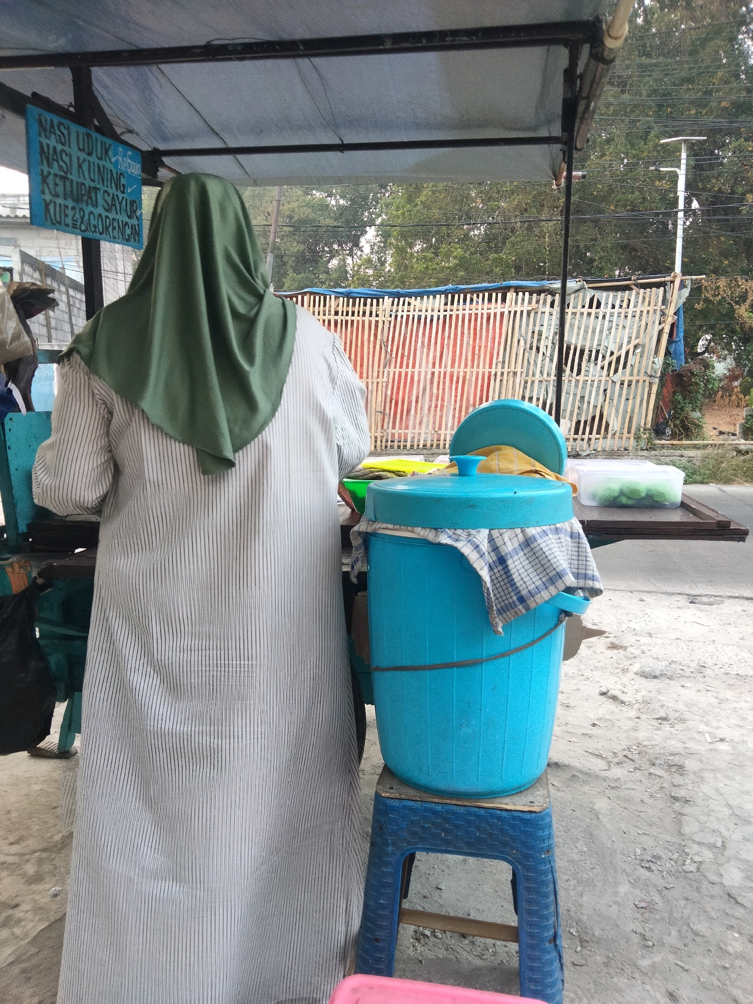 A person wearing a white pinstripe robe and a green hijab stands in front of a street food stall in Pisangan Jaya, Indonesia. The stall has a blue sign hanging from the roof with text that appears to be a menu. To the right of the person is a large blue bucket on a blue stool, covered with a checkered cloth. To the right of the bucket, on a wooden table, is a clear plastic container with green spherical objects inside. The background shows a bamboo fence and trees. The ground is made of gravel and concrete.