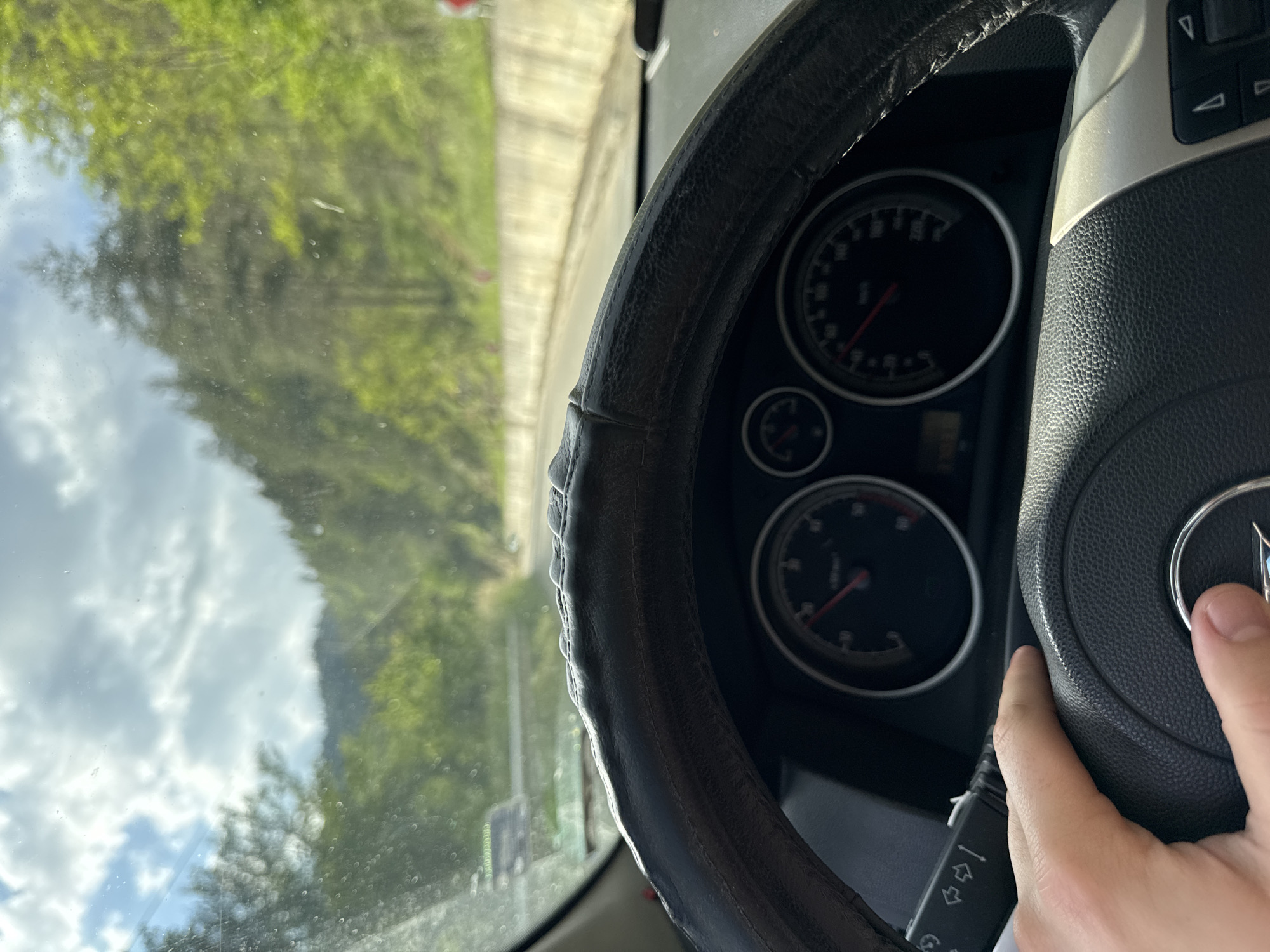 A car's interior is shown, with a view through the windshield of a road in Byala Reka, Bulgaria. The dashboard, steering wheel (with a worn cover), and instrument panel are visible. A hand is resting on the steering wheel. The exterior view features trees, sky with clouds, and a stretch of road.