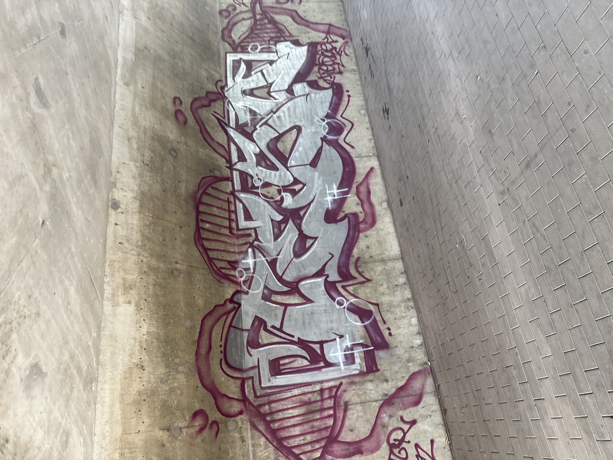 The image depicts a section of a concrete underpass in Rathenow, Germany, featuring extensive graffiti. The graffiti is primarily in shades of silver and deep pink, with intricate lettering and flowing designs covering a significant portion of the concrete wall. The style appears to be a complex graffiti piece with multiple layers and visual effects. To the left of the main graffiti, there are smaller pink markings, including what looks like a stylized letter 'R' and some abstract shapes. The ground beneath the underpass is paved with grey rectangular bricks arranged in a regular pattern. The lighting suggests it is daytime, and the overall scene conveys an urban, somewhat grimy atmosphere common to underpasses. There are no people visible in the image.