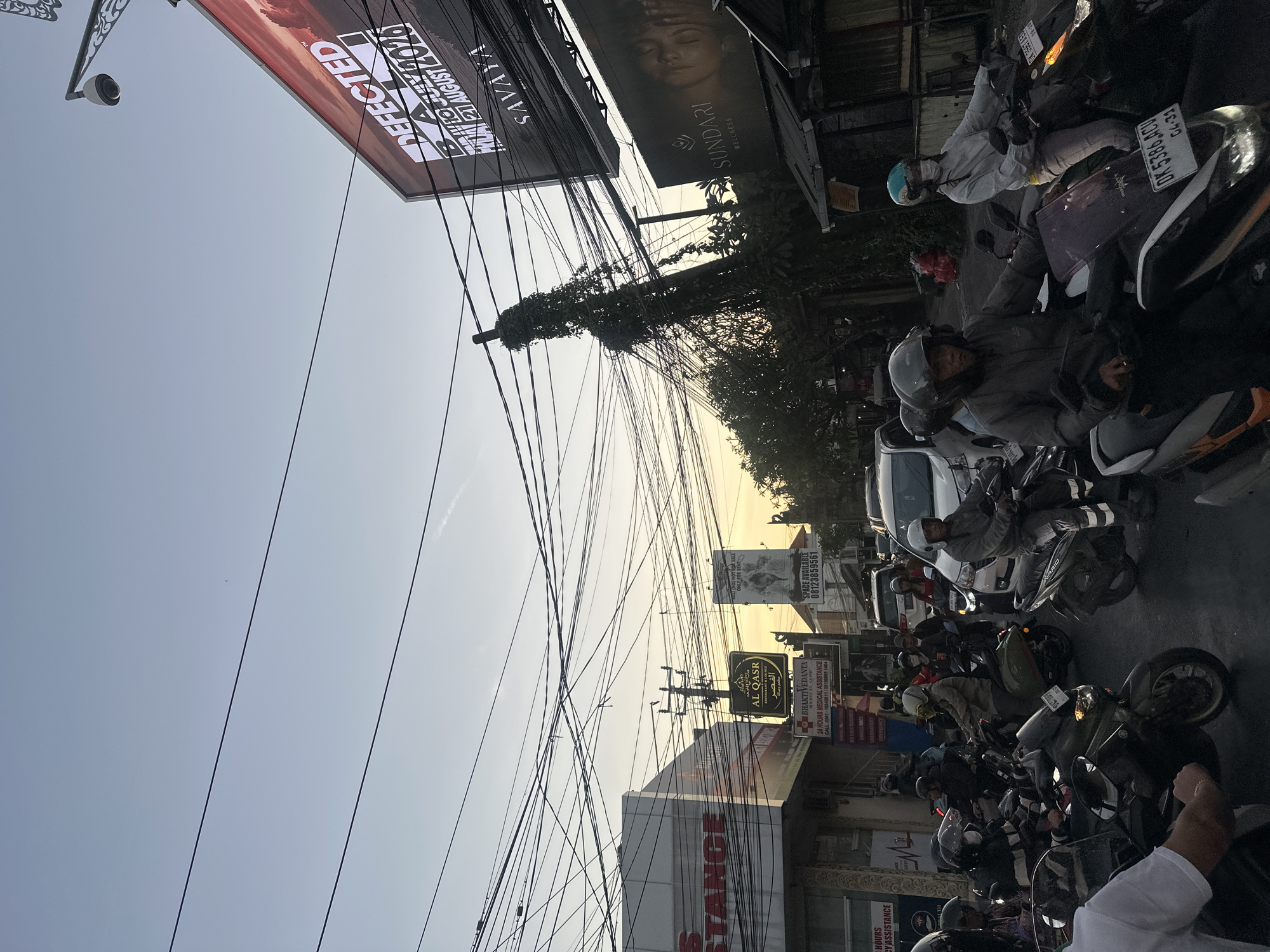 This media file depicts a busy street scene in North Kuta, Indonesia, likely during the late afternoon or early evening as indicated by the warm, low-angled sunlight and hazy sky. The foreground is dominated by a dense network of electrical wires, a common sight in many parts of Asia.
The street is filled with a variety of motorbikes, many with riders wearing helmets and jackets, suggesting a common mode of transportation and the need for protection. Some of the motorbikes are parked, while others appear to be in motion, contributing to the impression of a bustling urban environment. The presence of a white car also adds to the traffic density.
Several signs are visible along the street, offering clues about businesses and services. One prominent sign reads "AL QASR RESTAURANT & LOUNGE" with Arabic text. Another sign indicates "BHAKTIVEDANTA MEDICAL CLINIC" and advertises "24 HOURS MEDICAL ASSISTANCE." There is also a sign for "SUNDARI WELLNESS." A large billboard in the upper left corner features the word "DEFECTED" and a date, likely advertising an event. Another sign mentions "SPACE AVAILABLE" with a phone number, suggesting commercial activity.
The overall mood of the image is one of vibrant, everyday life, characterized by activity, commerce, and the characteristic urban infrastructure of the region. The sky is a soft blue, transitioning towards a warmer tone, hinting at the approaching sunset.