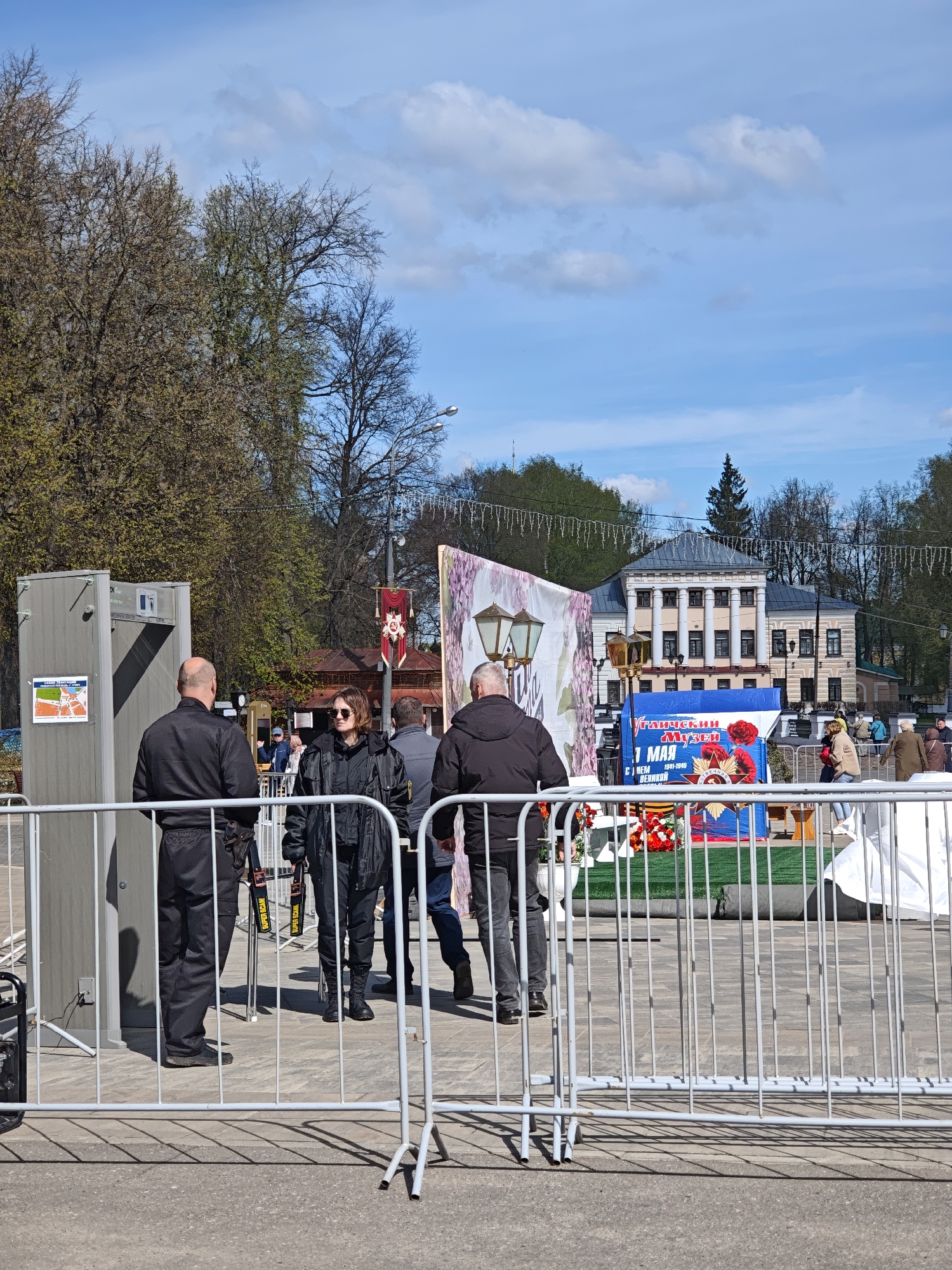 A daytime image shows a security checkpoint at what appears to be an event entrance in Uglich, Russia. Visible is a metal detector, operated by a man in a black uniform, and a fenced-off area. A woman in a black jacket and sunglasses is walking toward the checkpoint. Other people can be seen passing through the area. A blue banner with Russian text and a star emblem is visible, along with decorations including flowers. In the background, a building with columns can be seen. The environment includes trees and a partially cloudy sky.