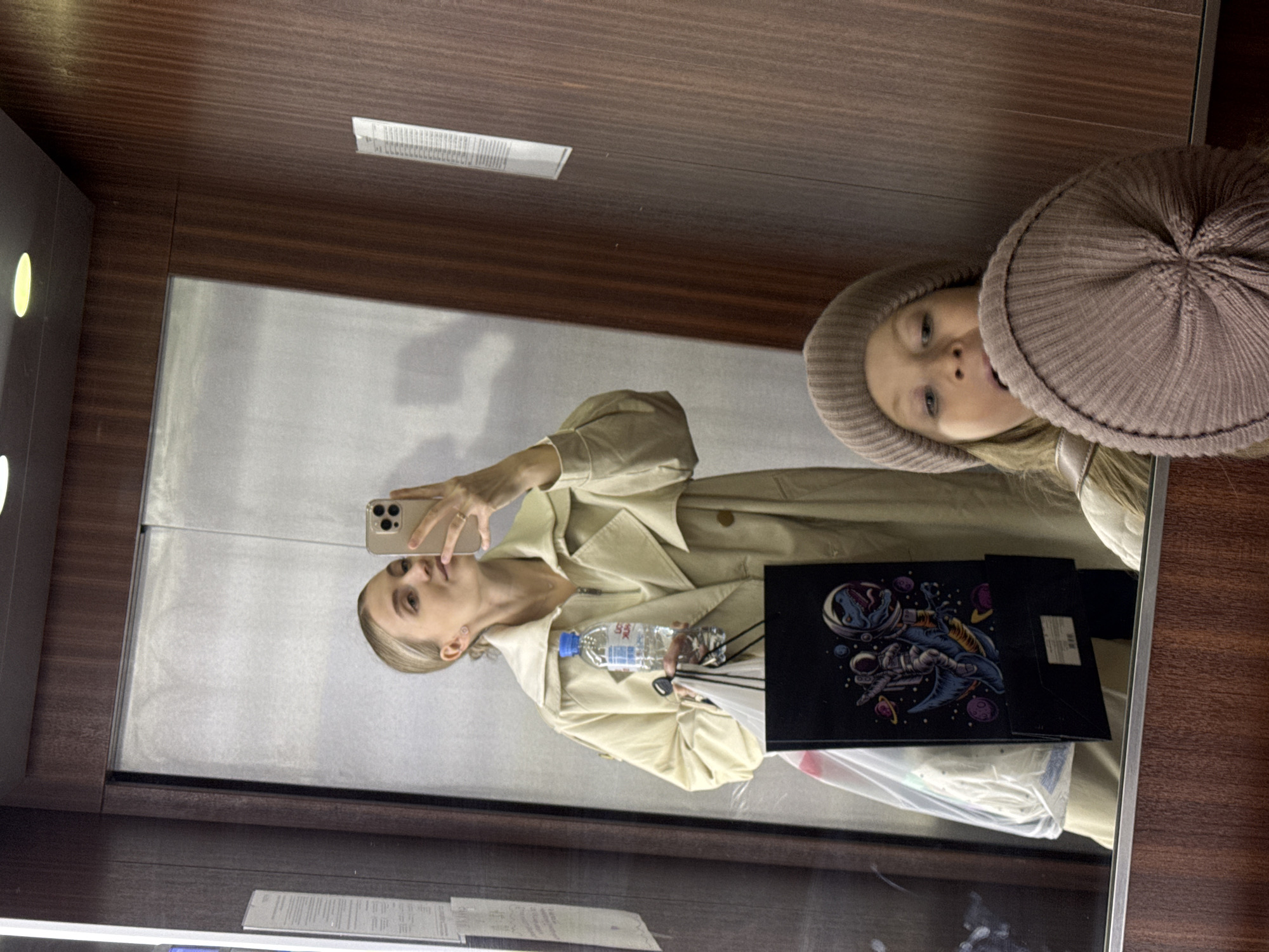 A mirror selfie shows a woman wearing a beige trench coat and a brown beanie. She holds a water bottle in one hand and a black bag with a space-themed graphic. The reflection suggests she is in an elevator with wood-paneled walls. The location is unknown and cannot be reliably determined to be in the city of Izhevsk, Russia, from this image alone.