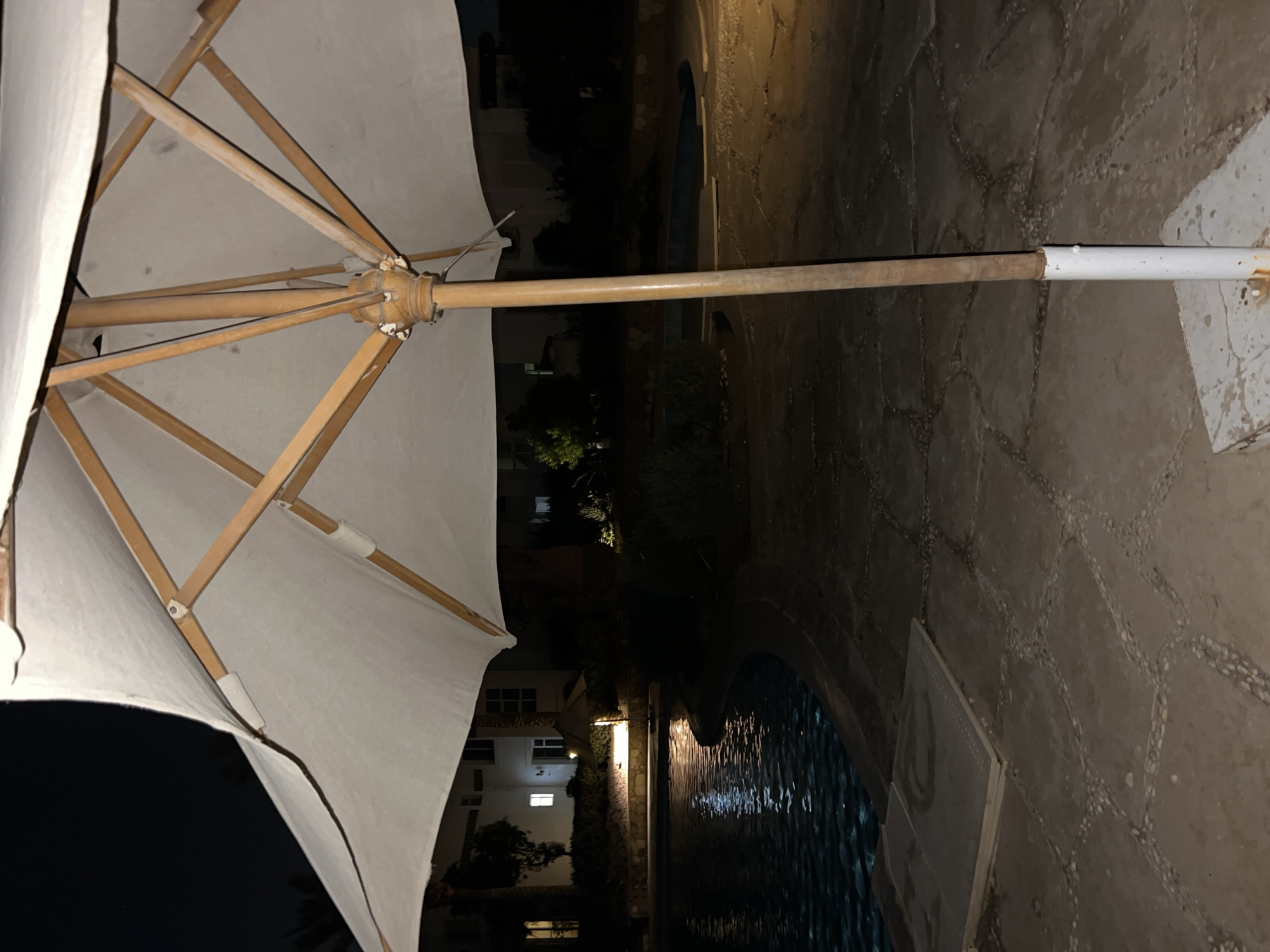 The image depicts an outdoor scene at night, likely in Sharm El Sheikh, Egypt, with a focus on a large white patio umbrella in the foreground. The umbrella is partially open and angled, revealing its wooden ribs and central pole. The pole is secured by a white metal casing at its base, which is set in a concrete block. To the left of the umbrella, a swimming pool with gently rippling, dark water is visible, reflecting ambient light from nearby buildings. The pool area is paved with irregular-shaped stones, interspersed with smaller pebbles. In the background, several multi-story buildings are illuminated, suggesting a resort or residential complex. Some vegetation, including palm trees, is present between the pool and the buildings. The overall atmosphere is dark and quiet, characteristic of an evening setting in a warm climate. There are no people or animals visible in the frame.