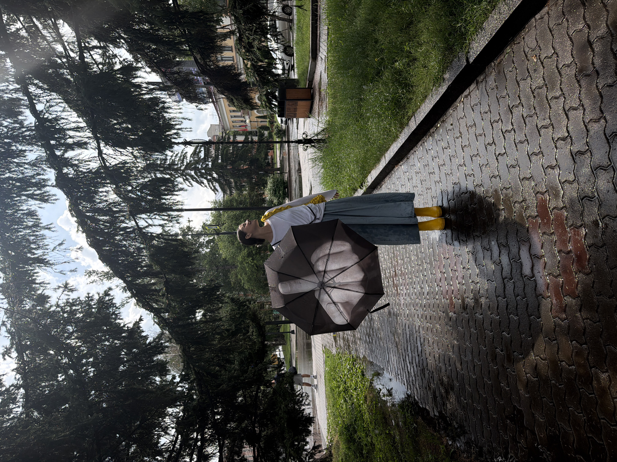 In a park-like setting in the city of Krasnoyarsk, Russia, a woman stands on a wet, brick-paved walkway under a cloudy sky. She is holding a closed umbrella with a graphic of a hand giving the middle finger. The woman is wearing a white long-sleeved shirt, a blue skirt, and yellow rain boots. To her left, a man walks away from the camera on the same wet walkway. Lush green grass lines the right side of the walkway, and large pine trees with drooping branches overhang the scene, casting shadows and creating a sense of enclosure. In the background, buildings and parked cars suggest an urban environment bordering the park. The wet pavement indicates recent or ongoing rain.