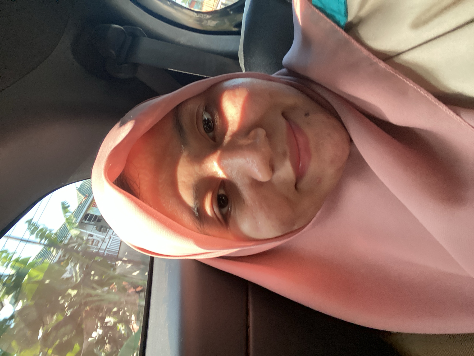 A woman is taking a selfie in the back seat of a car. She is wearing a pink hijab and a beige top with a teal collar. The sun is shining through the window, casting shadows on her face. Outside the window, there are trees and a building. The car appears to be moving. The location is specified as Antasan Senor Ilir, Indonesia, though this information is not visually verifiable from the image itself.